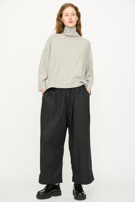 A person stands against a plain background wearing the Ichi Antiquités Cotton Turtleneck in Gray, wide black pants, and black shoes, with hands in pockets.