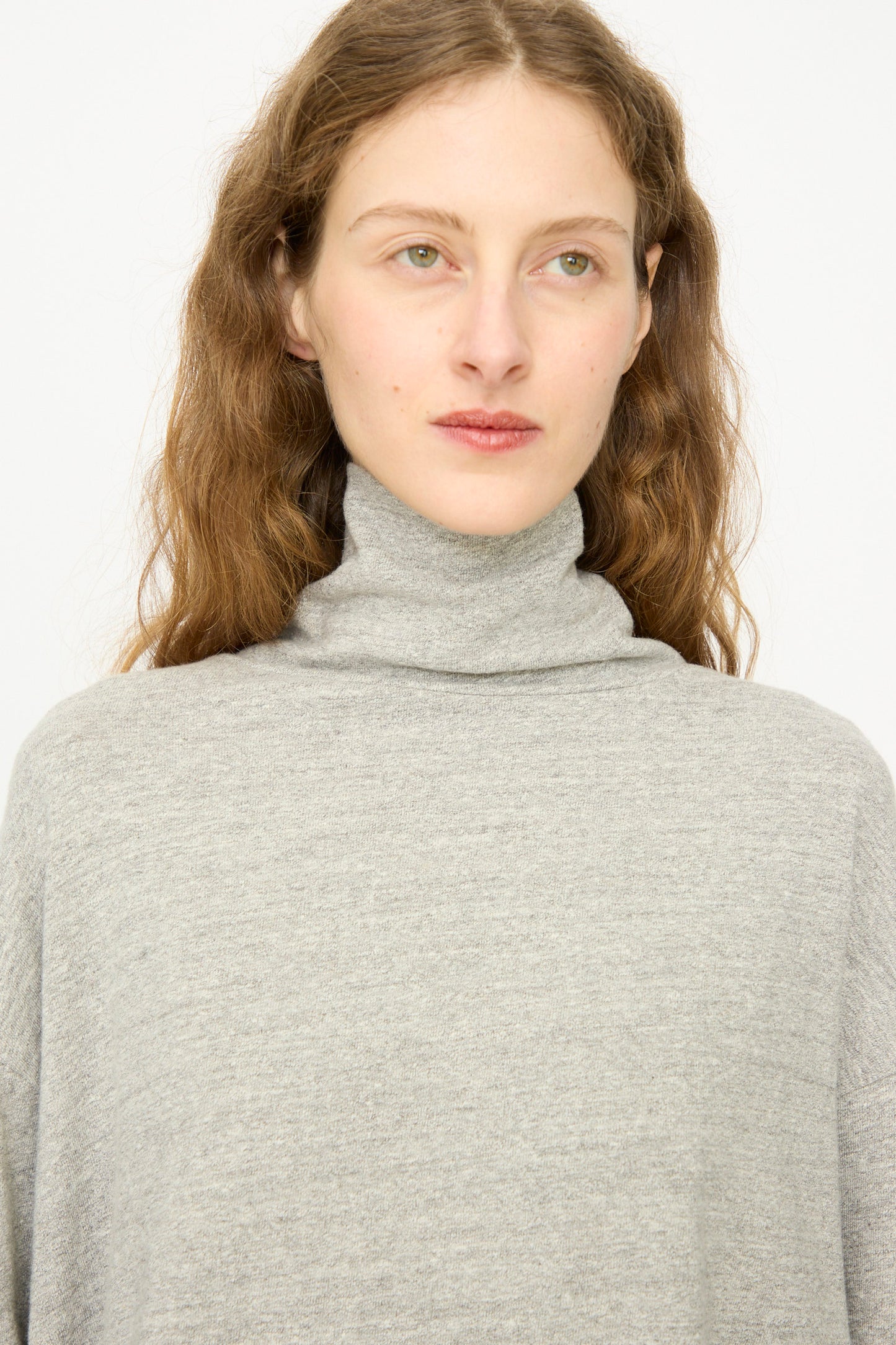 A person with long, wavy brown hair wears the Ichi Antiquités Cotton Turtleneck in Gray, standing against a plain white background.