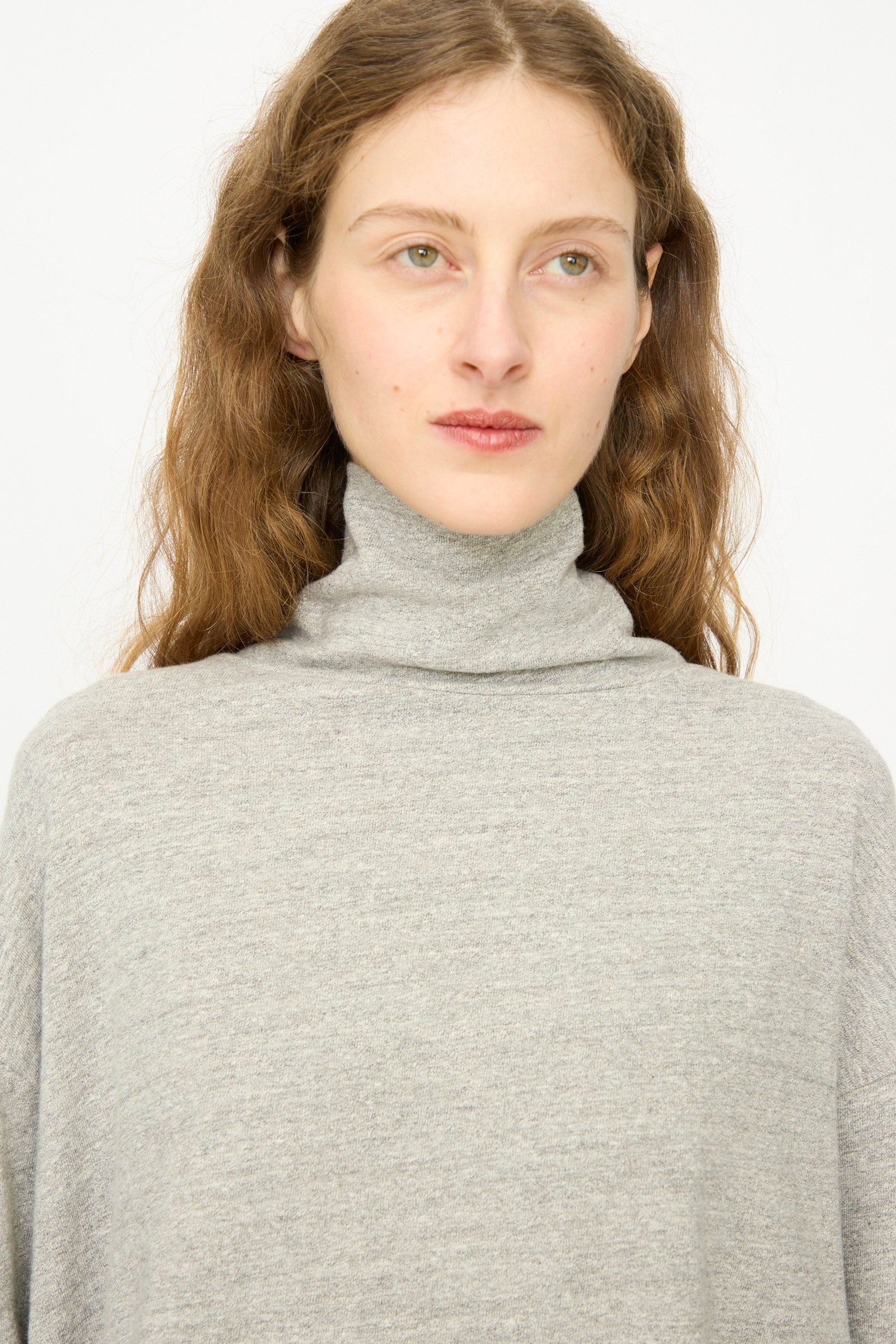 A person with long, wavy brown hair wears the Ichi Antiquités Cotton Turtleneck in Gray, standing against a plain white background.