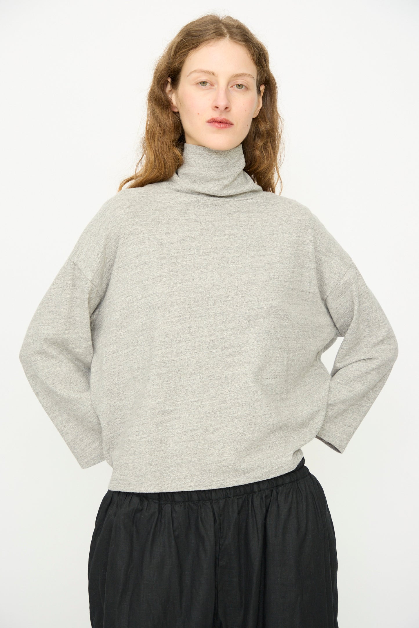 A person with long brown hair models the Ichi Antiquités Cotton Turtleneck in Gray, paired with loose black pants, against a plain white background.