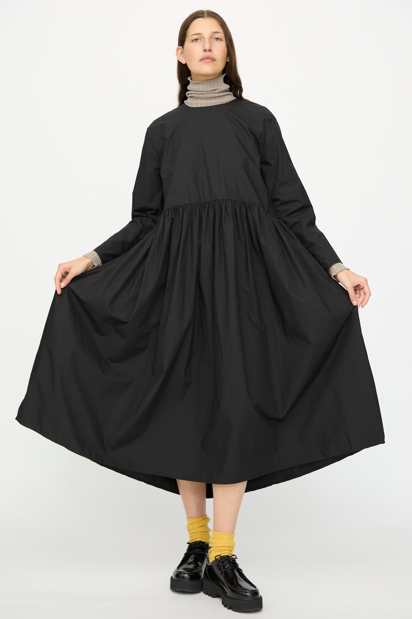 A person wearing the Ichi Antiquités Cotton Typewriter Dress in Black, layered over a gray turtleneck with yellow socks and black shoes, stands against a plain white background.