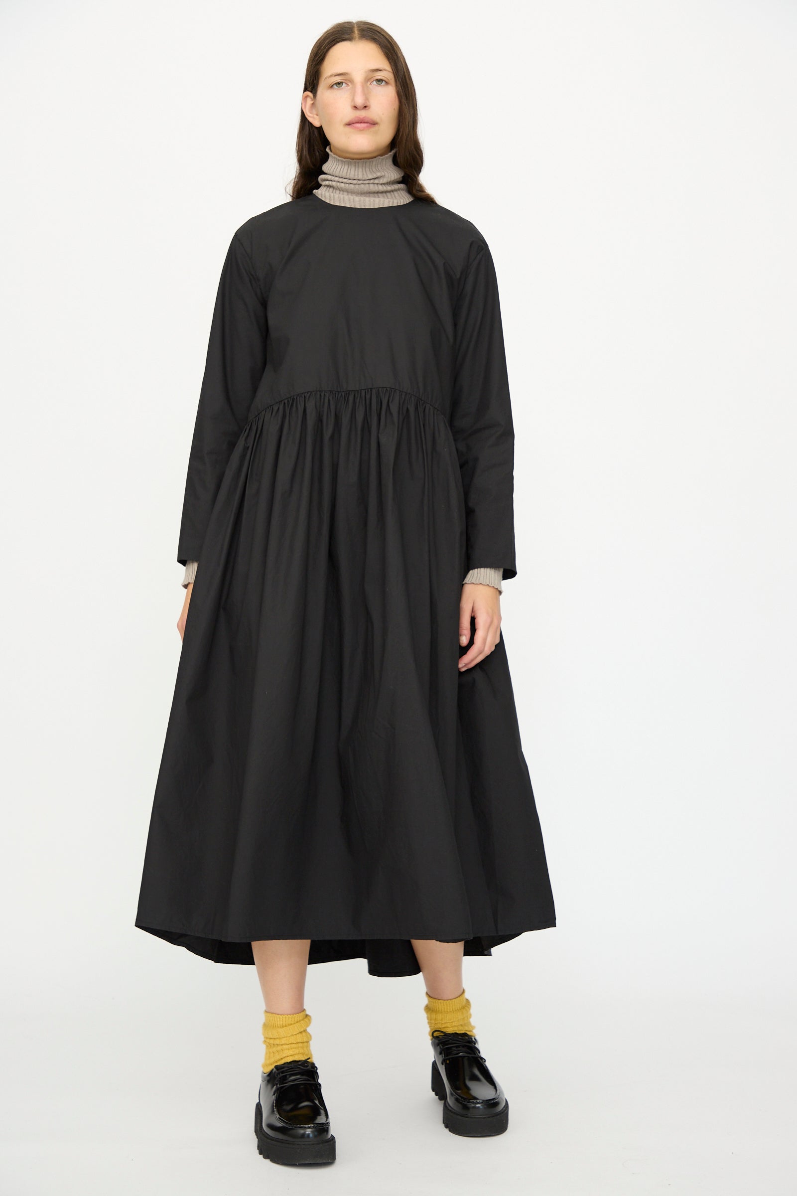 A person wears the Ichi Antiquités Cotton Typewriter Dress in Black over a beige turtleneck, styled with yellow socks and black shoes, standing against a plain background.