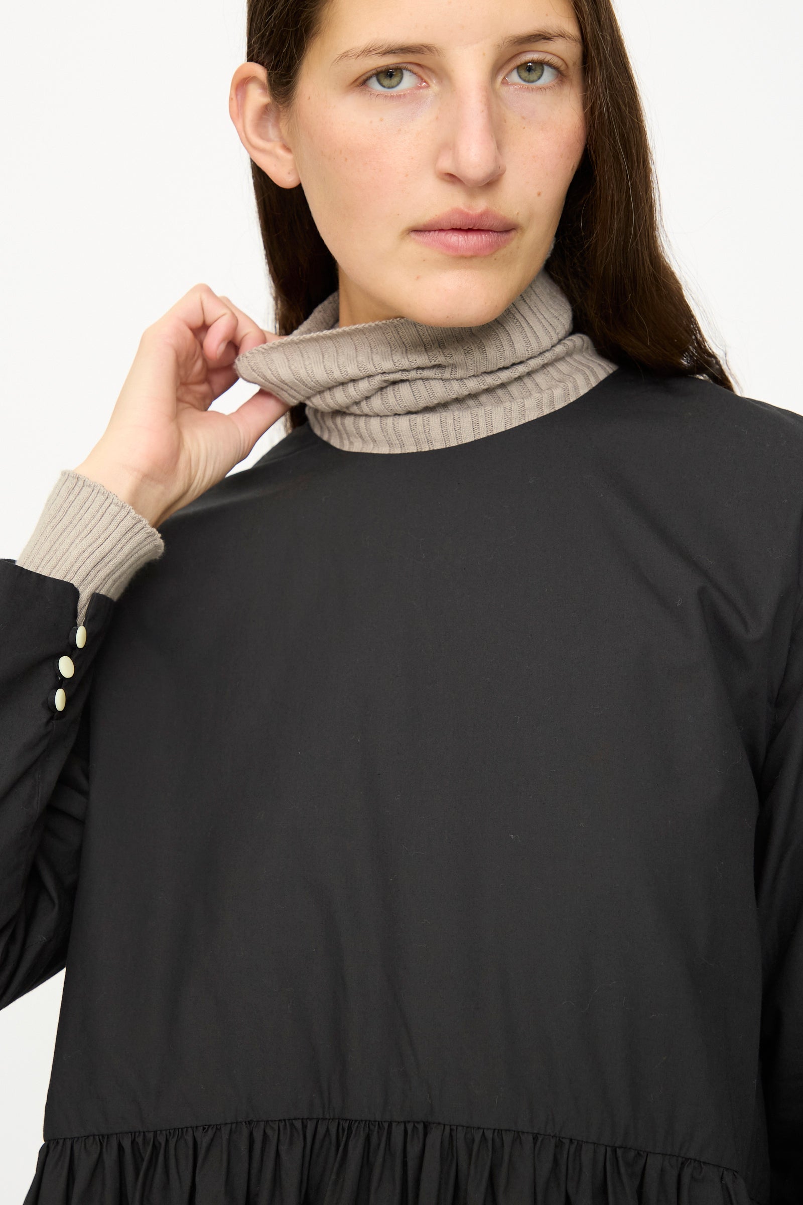 Wearing the Ichi Antiquités Cotton Typewriter Dress in Black over a beige turtleneck, a person with long brown hair holds the collar and looks at the camera.