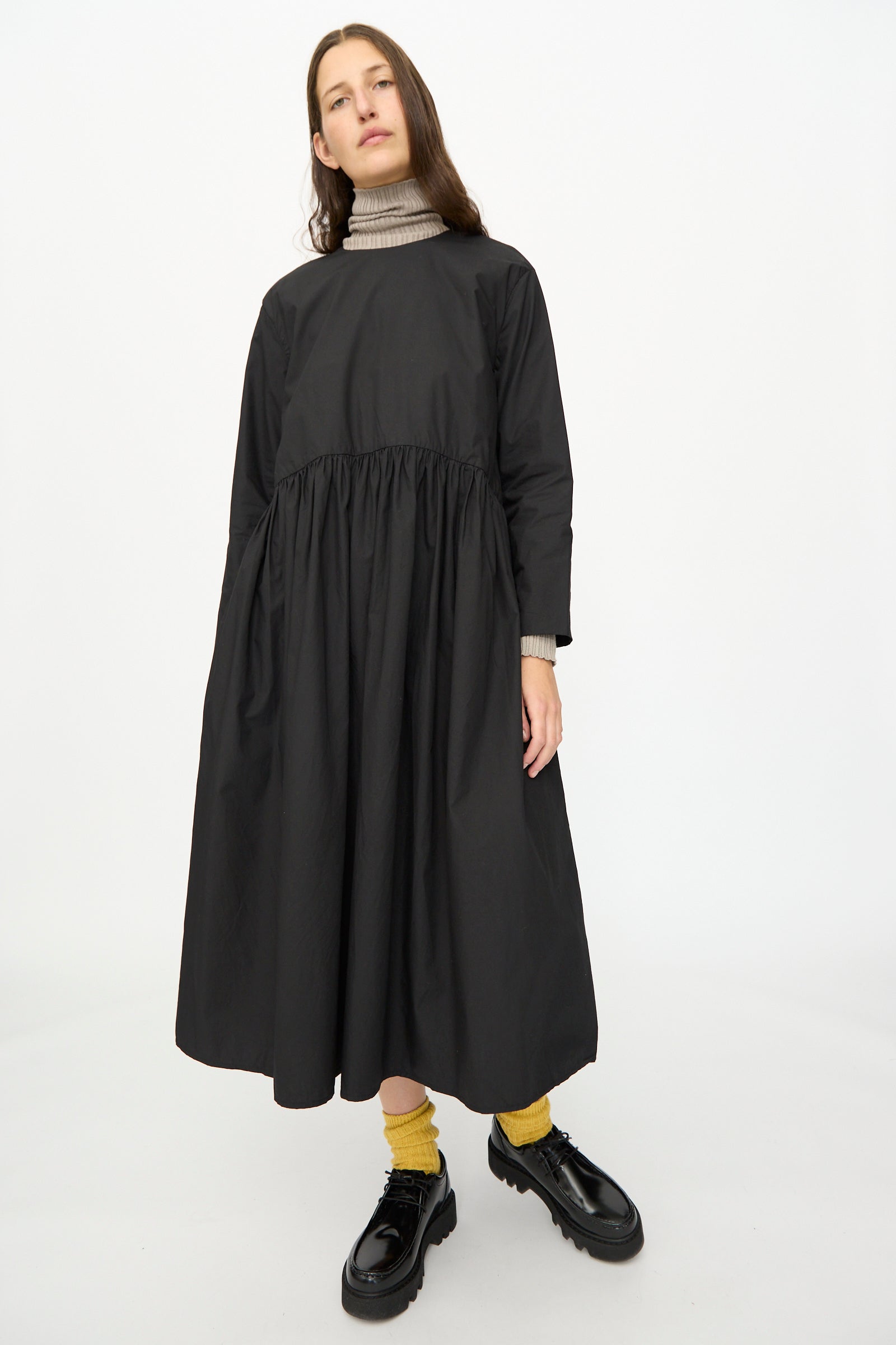 A person models the Ichi Antiquités Cotton Typewriter Dress in Black over a beige turtleneck, paired with yellow socks and black shoes, against a plain white background.