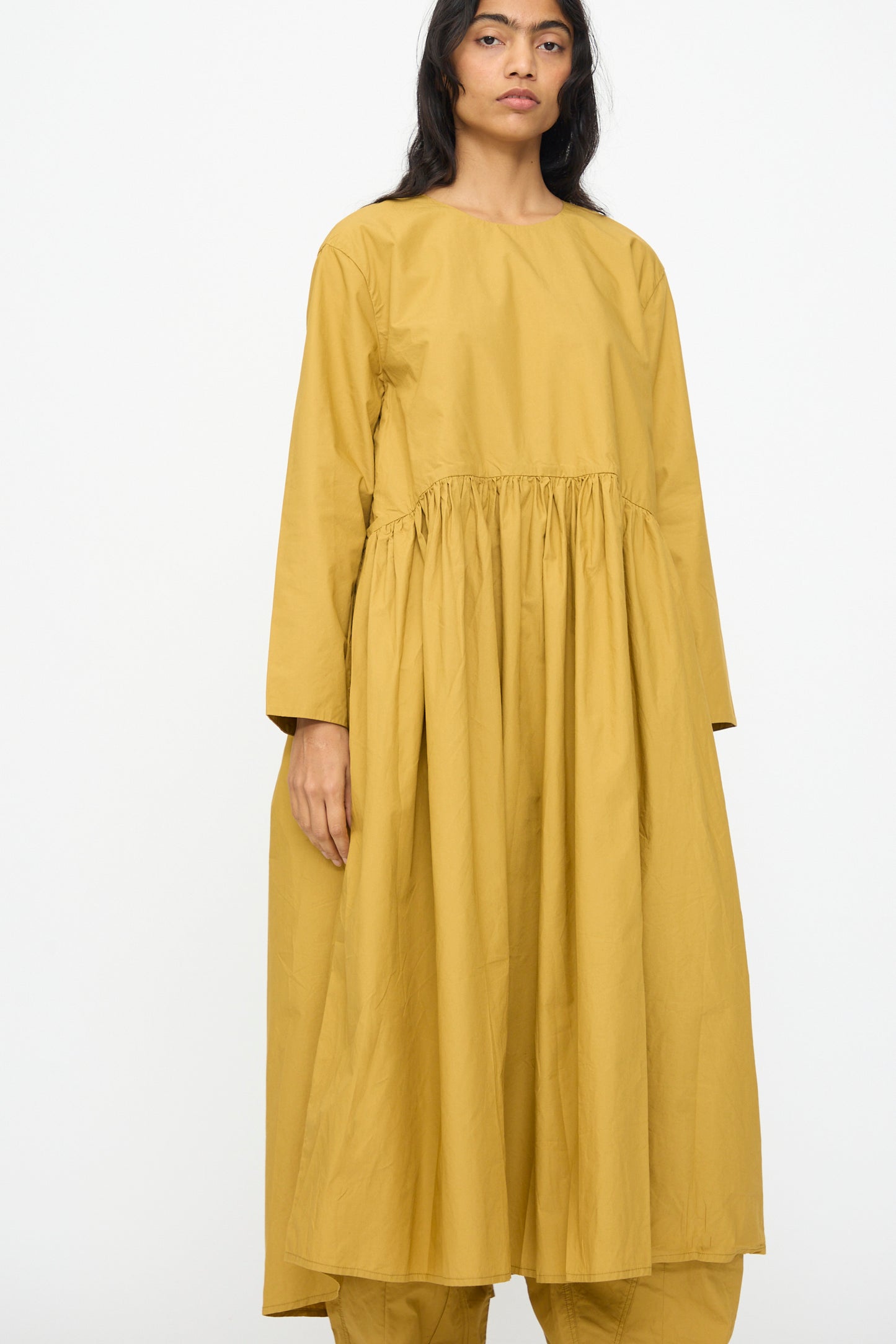 A model wears the Ichi Antiquités Cotton Typewriter Dress in mustard, featuring long sleeves, an oversized fit, and a gathered waist, standing against a plain white background.