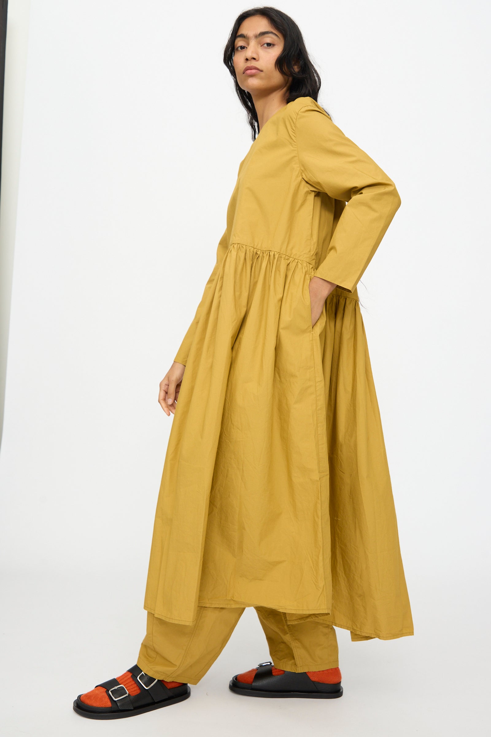 A person in the Ichi Antiquités Cotton Typewriter Dress in Mustard, paired with matching pants, black sandals, and orange socks, stands against a plain white background.