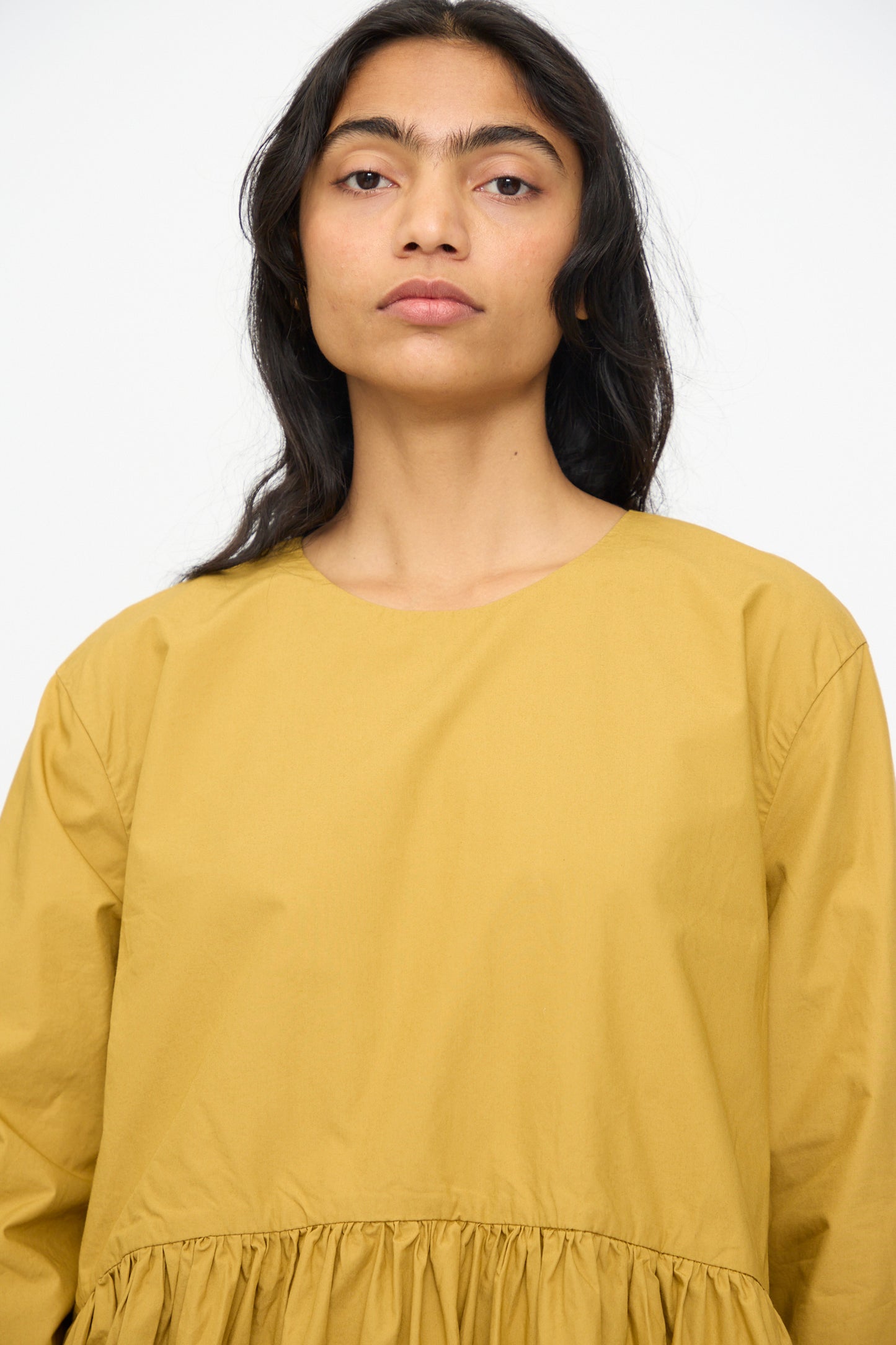 A person with long dark hair models the Ichi Antiquités Cotton Typewriter Dress in Mustard, a loose-fitting cotton dress with a gathered waist, set against a plain white background.