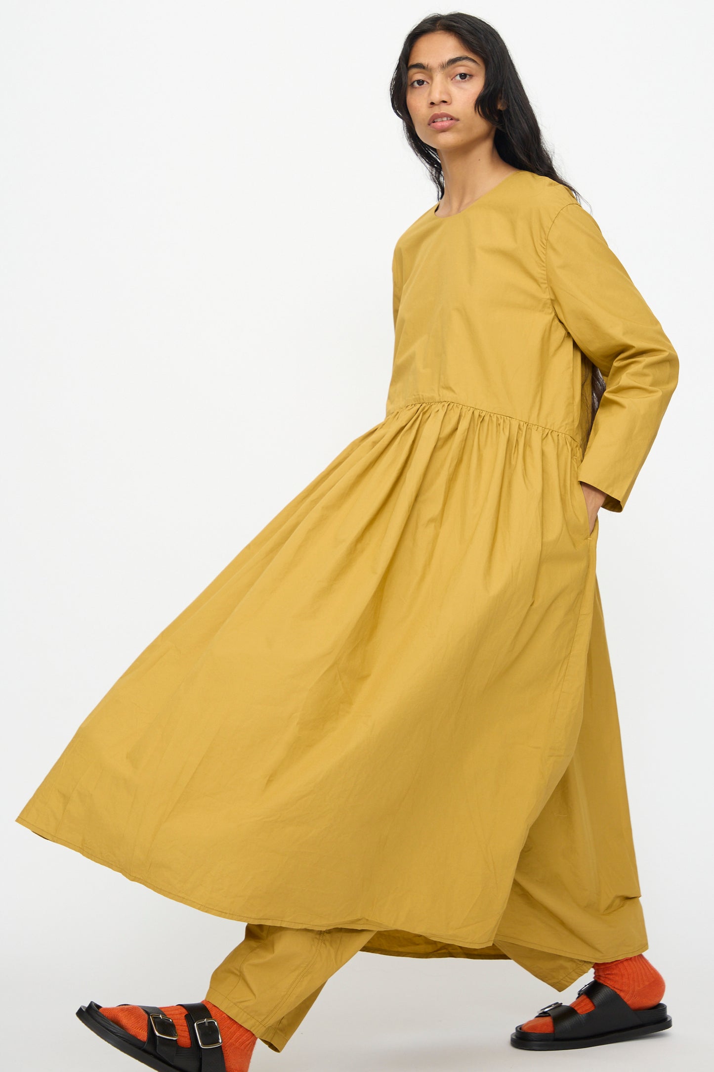 A person wears the Ichi Antiquités Cotton Typewriter Dress in Mustard, styled oversized over matching pants, with orange socks and black sandals, walking against a plain white background.