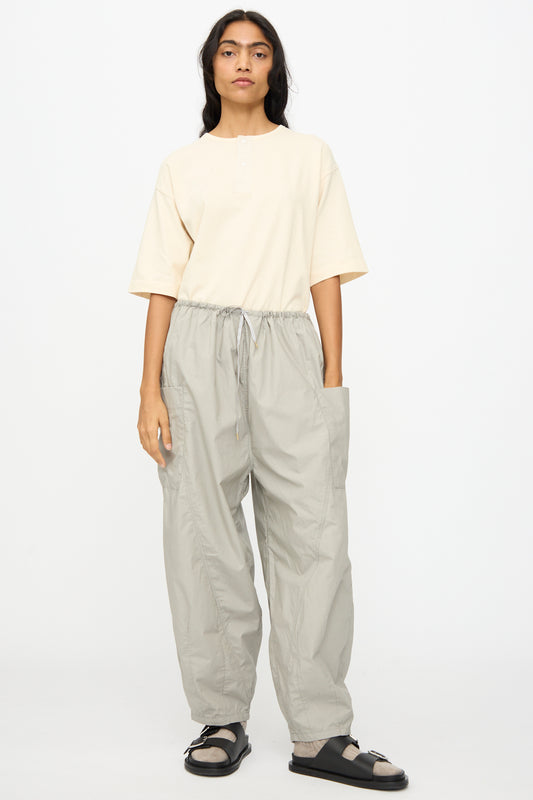 A person stands against a plain background wearing gray Cotton Typewriter Pants by Ichi Antiquités, paired with a light beige short-sleeve shirt and black sandals with white socks.