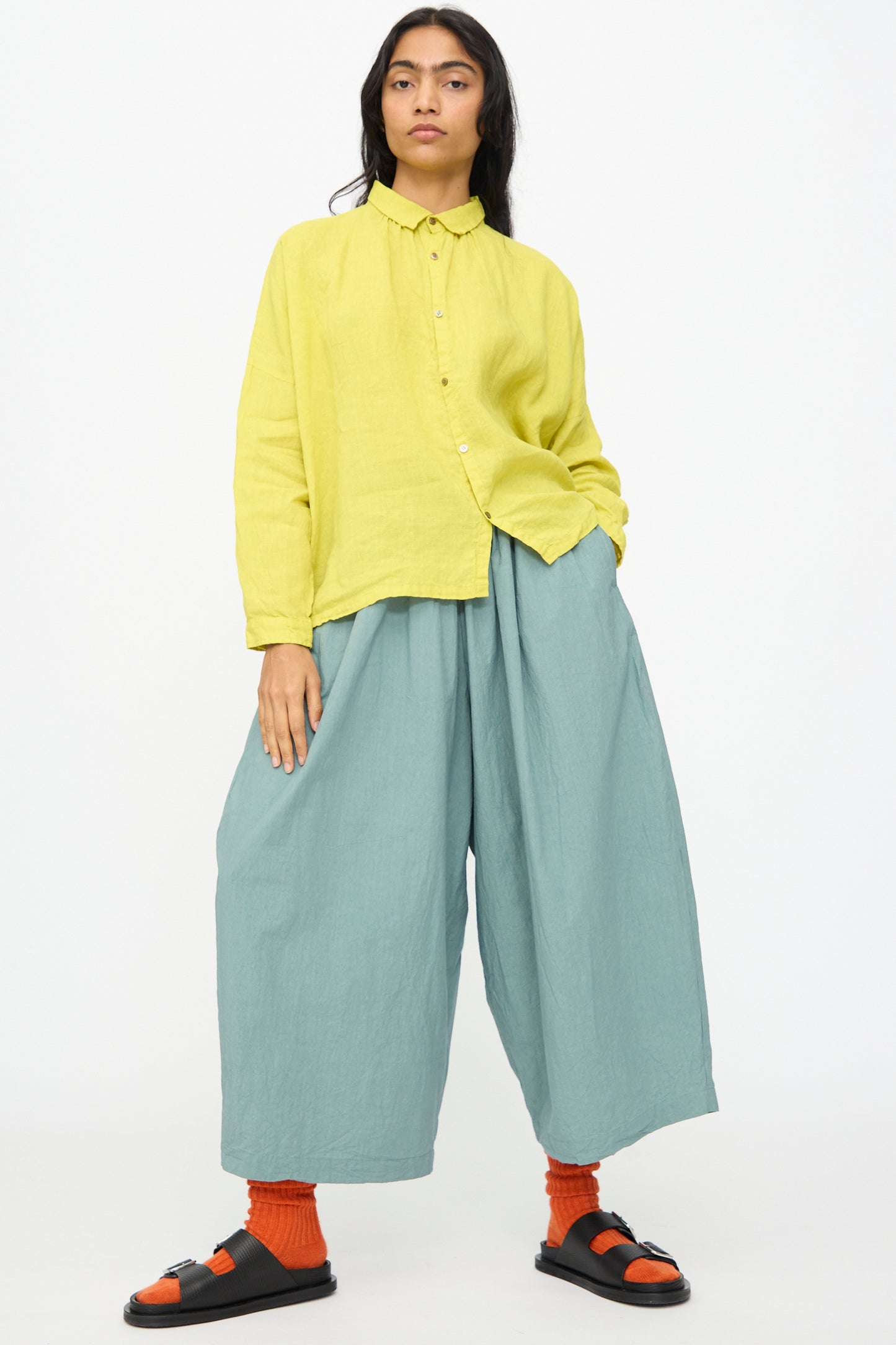 A person wears a loose yellow button-up shirt, Ichi Antiquités Cotton Typewriter Pants in Ice Blue, orange socks, and black sandals, standing against a plain white background.