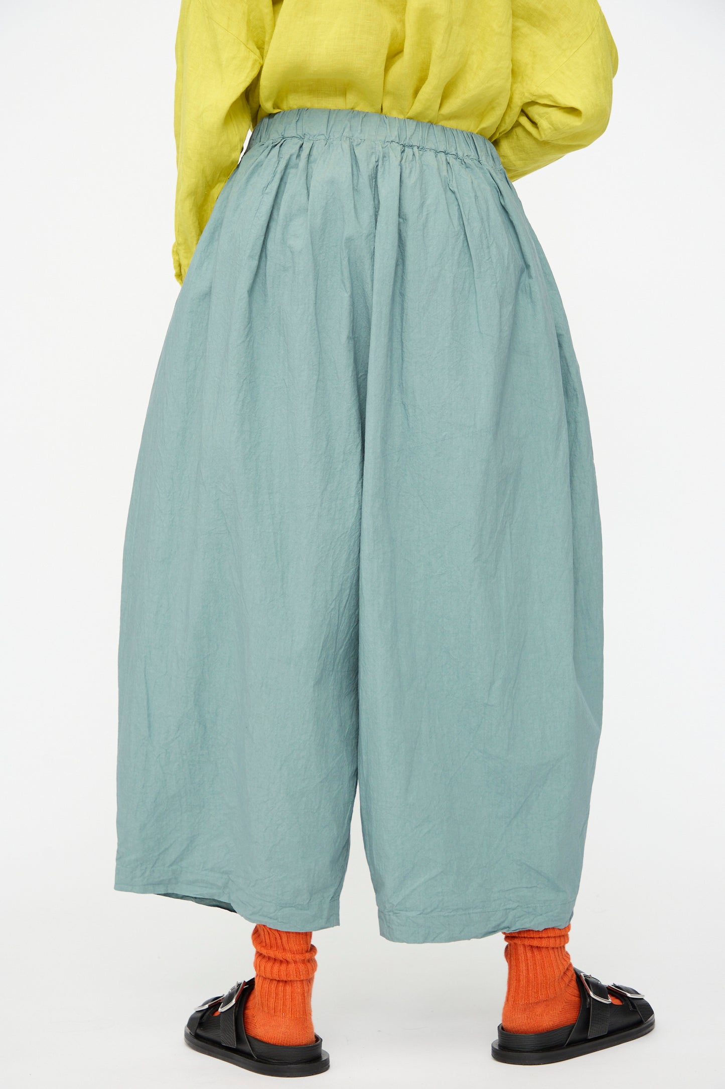 Person wearing Ichi Antiquités Cotton Typewriter Pants in Ice Blue, paired with a yellow top, orange socks, and black sandals, standing against a plain white background and shown from the back.