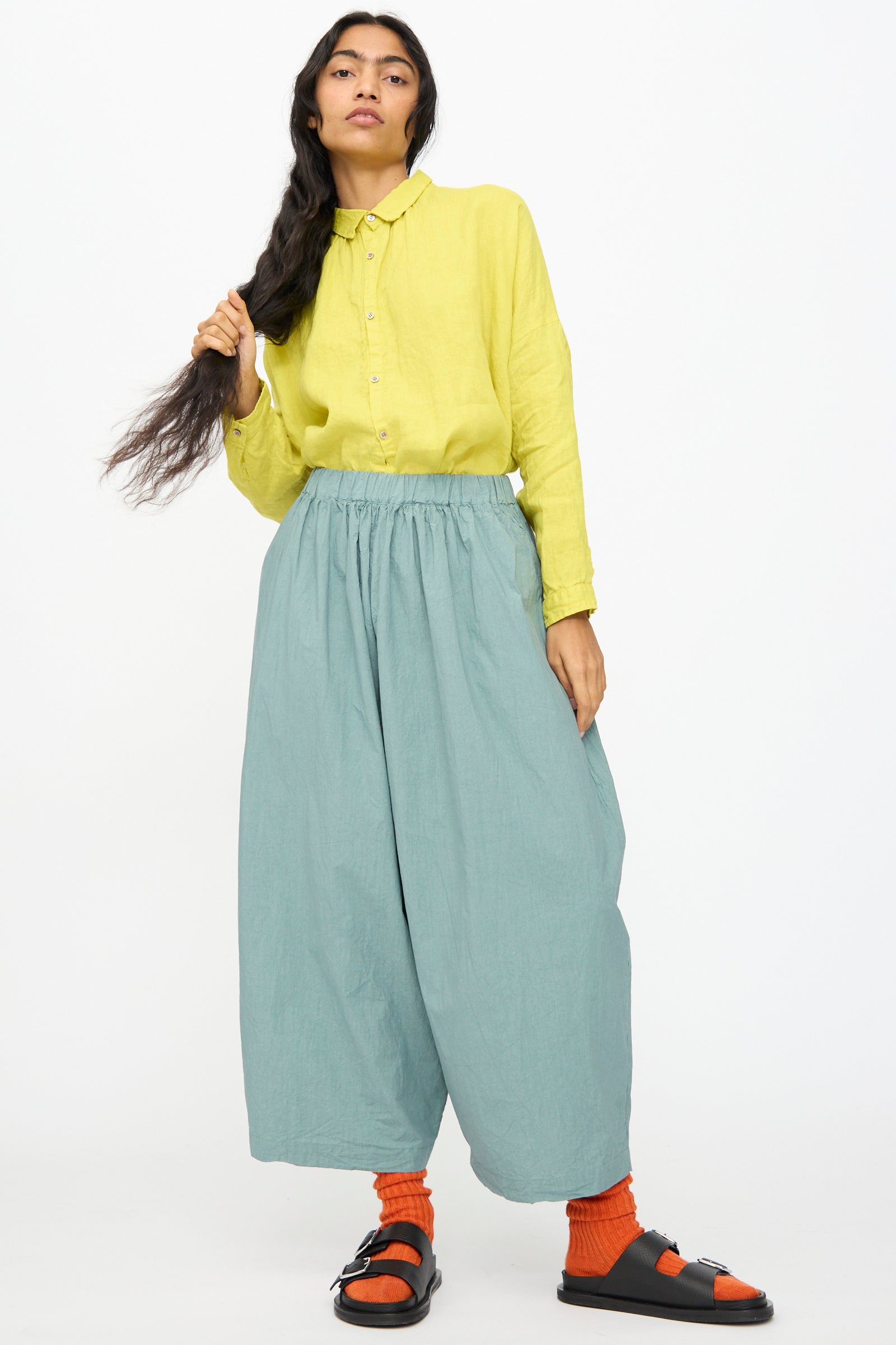 A person stands against a plain background wearing Ichi Antiquités Cotton Typewriter Pants in Ice Blue, styled with a yellow button-up shirt, bright orange socks, and black sandals.