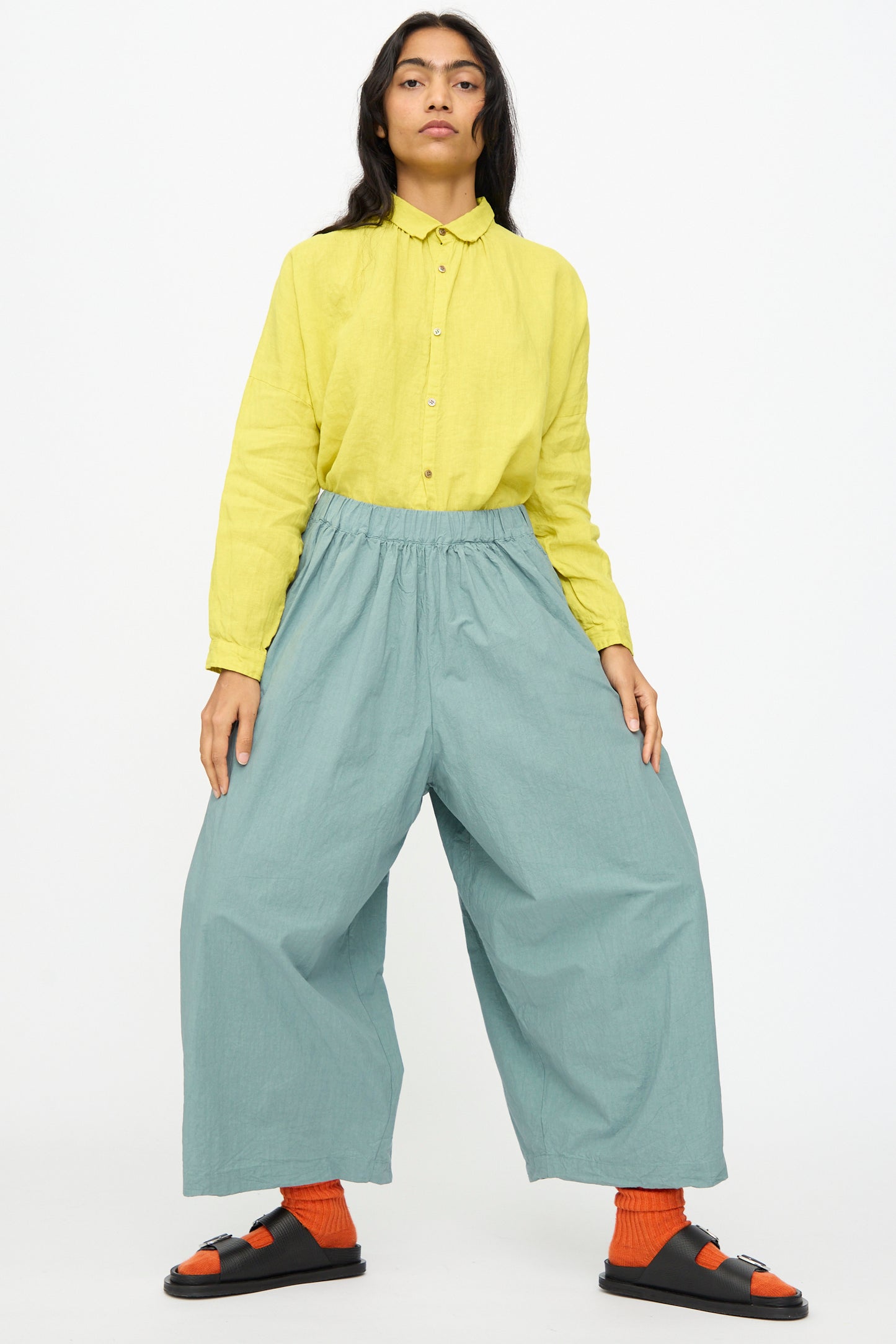 A person wears Ichi Antiquités' Cotton Typewriter Pants in Ice Blue with a yellow shirt, orange socks, and black sandals, standing against a plain light background.