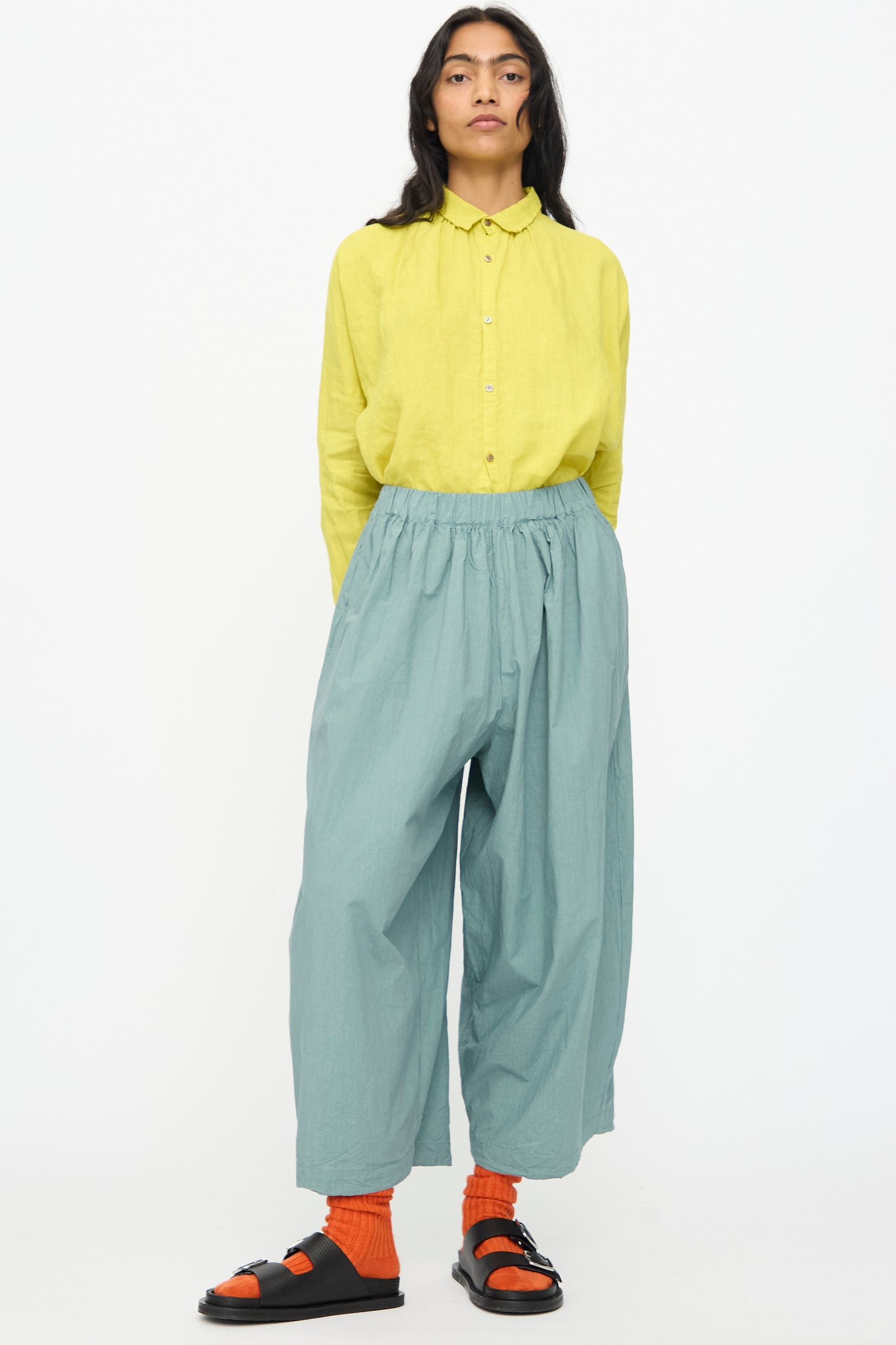 Person standing against a white background, wearing a yellow long-sleeve shirt, orange socks, black sandals, and Ichi Antiquités Cotton Typewriter Pants in Ice Blue.