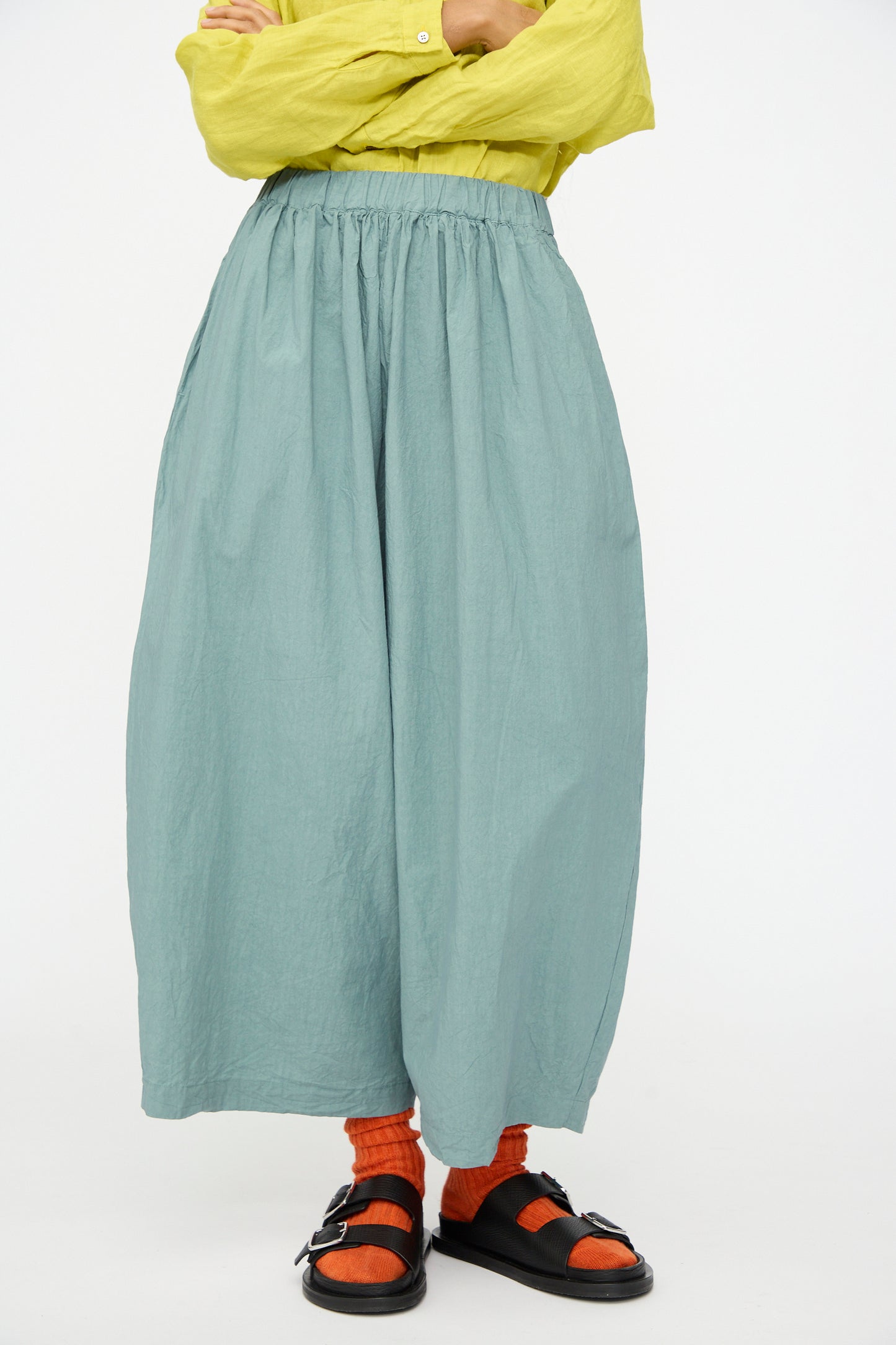 A person, face not visible, stands against a plain white background wearing a light green long-sleeve top, Ichi Antiquités Cotton Typewriter Pants in Ice Blue, orange socks, and black sandals.