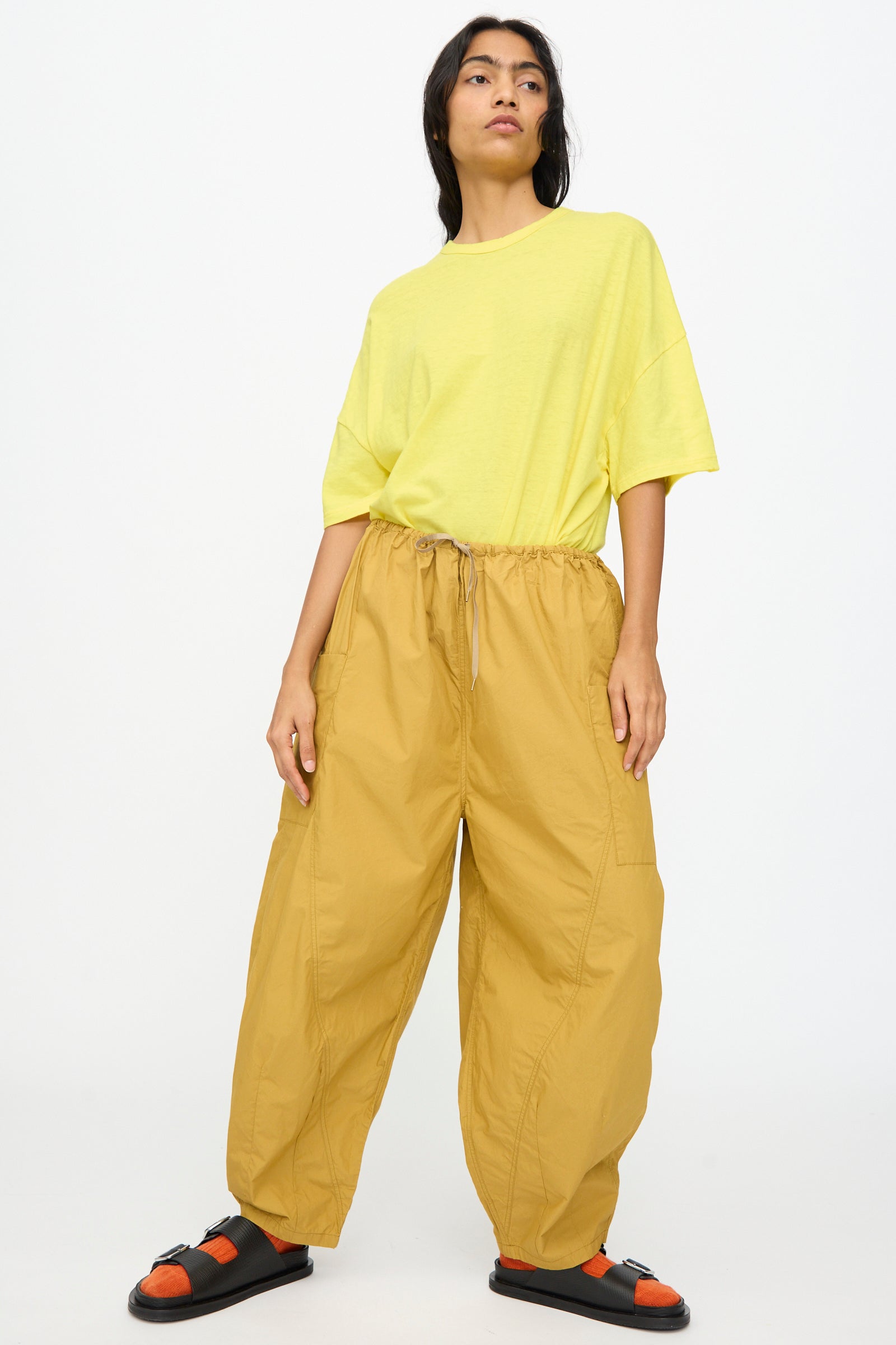 A person stands against a plain backdrop wearing a loose yellow t-shirt, Ichi Antiquités Cotton Typewriter Pants in Mustard with an elastic waist made in Japan, and black sandals with orange straps.