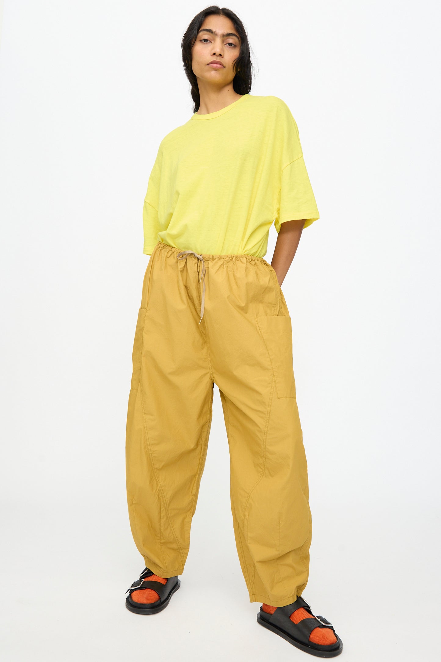 A person stands against a plain white background wearing Ichi Antiquités Cotton Typewriter Pants in Mustard, a loose yellow T-shirt, and black sandals with orange socks.