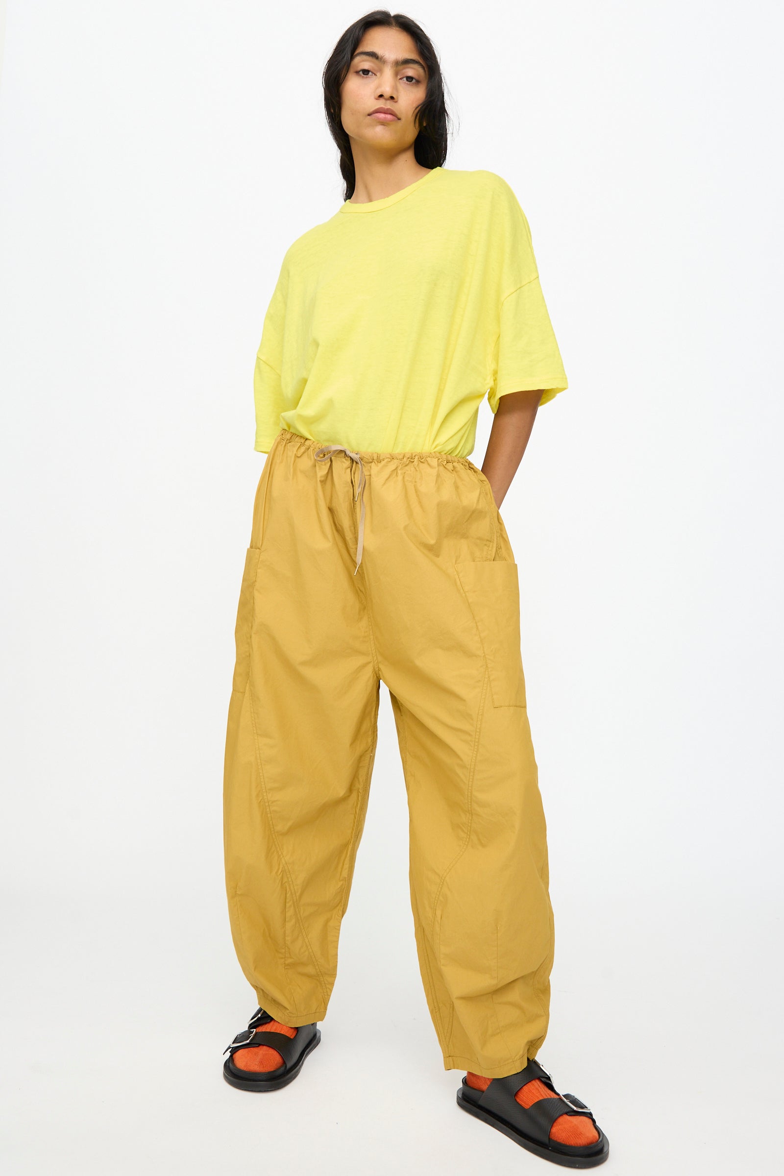 A person stands against a plain white background wearing Ichi Antiquités Cotton Typewriter Pants in Mustard, a loose yellow T-shirt, and black sandals with orange socks.