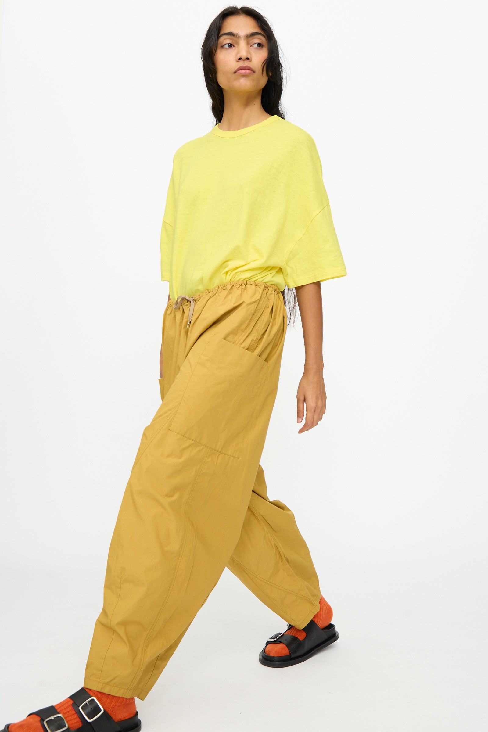 A person stands against a plain white background wearing Ichi Antiquités Cotton Typewriter Pants in Mustard, a loose yellow T-shirt, and black sandals with orange socks.