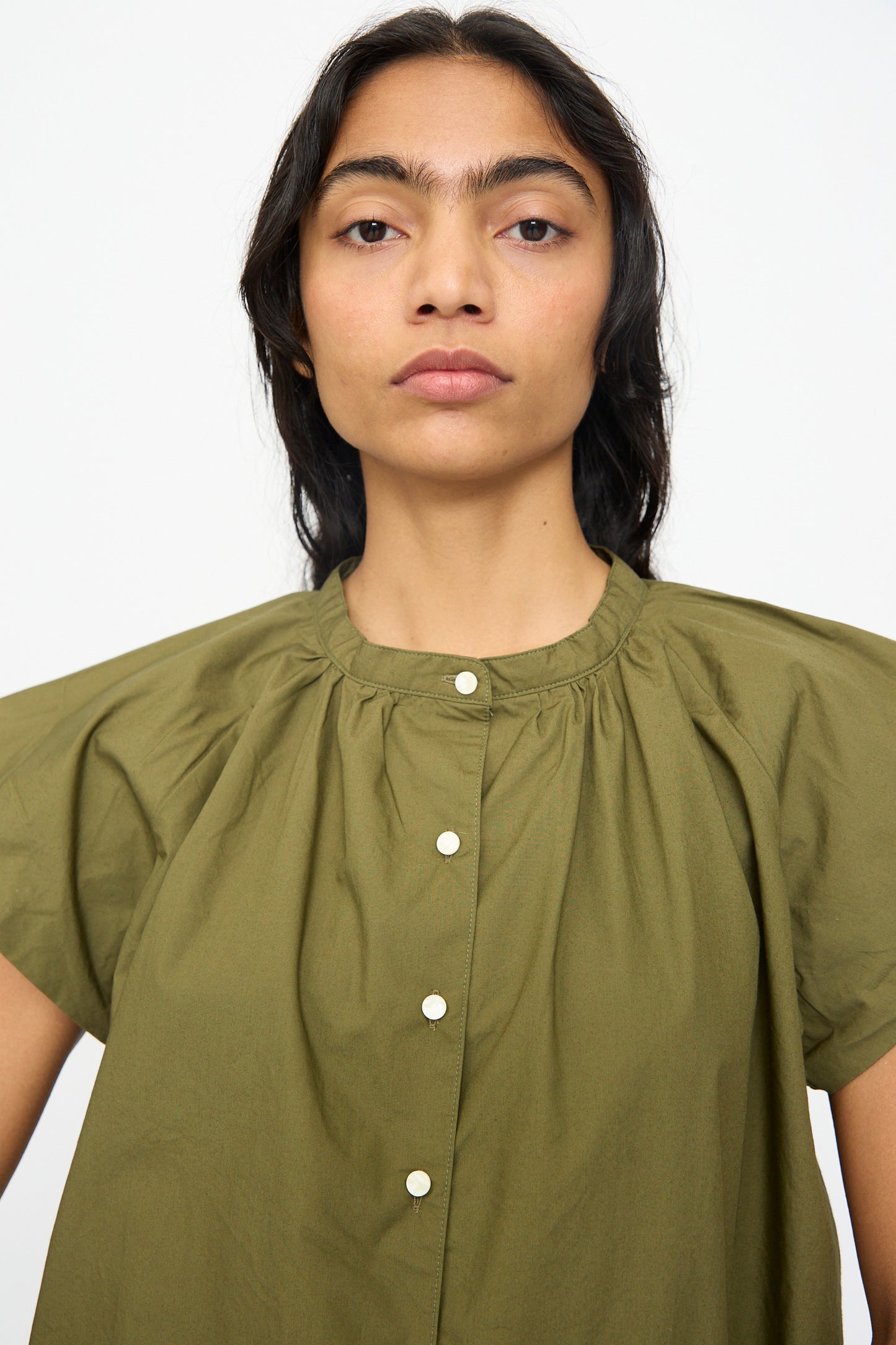A person with long dark hair wears the Ichi Antiquités Cotton Typewriter Shirt in Khaki, featuring short wide sleeves and front buttons, standing against a plain white background.