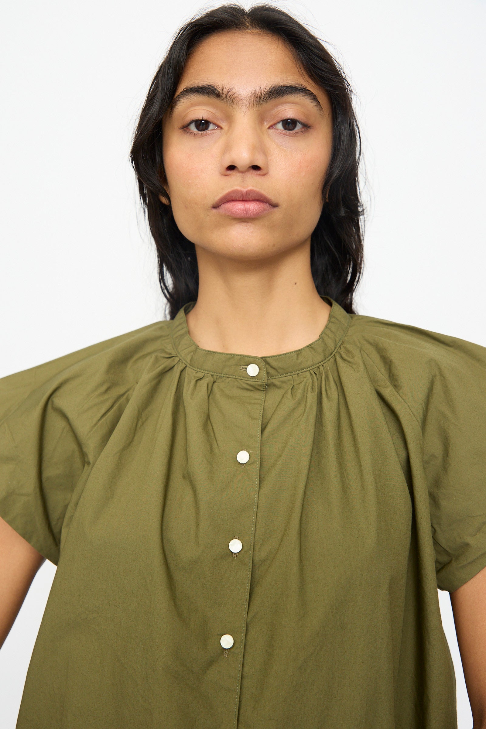 A person with long dark hair wears the Ichi Antiquités Cotton Typewriter Shirt in Khaki, featuring short wide sleeves and front buttons, standing against a plain white background.