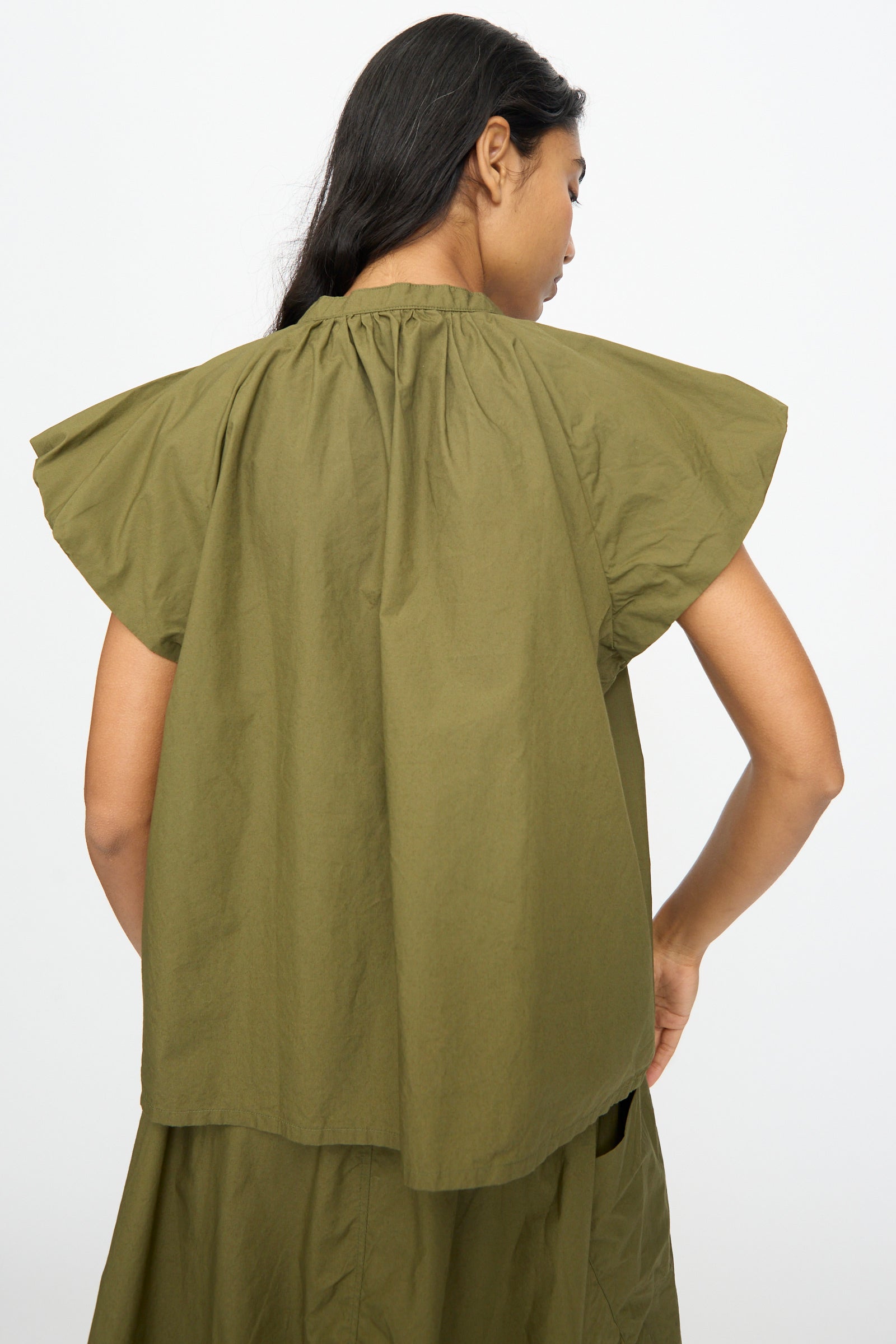 A woman with long dark hair, seen from the back, wears the Ichi Antiquités Cotton Typewriter Shirt in Khaki—an olive green, short-sleeve, loose-fitting cotton top with a gathered neckline.
