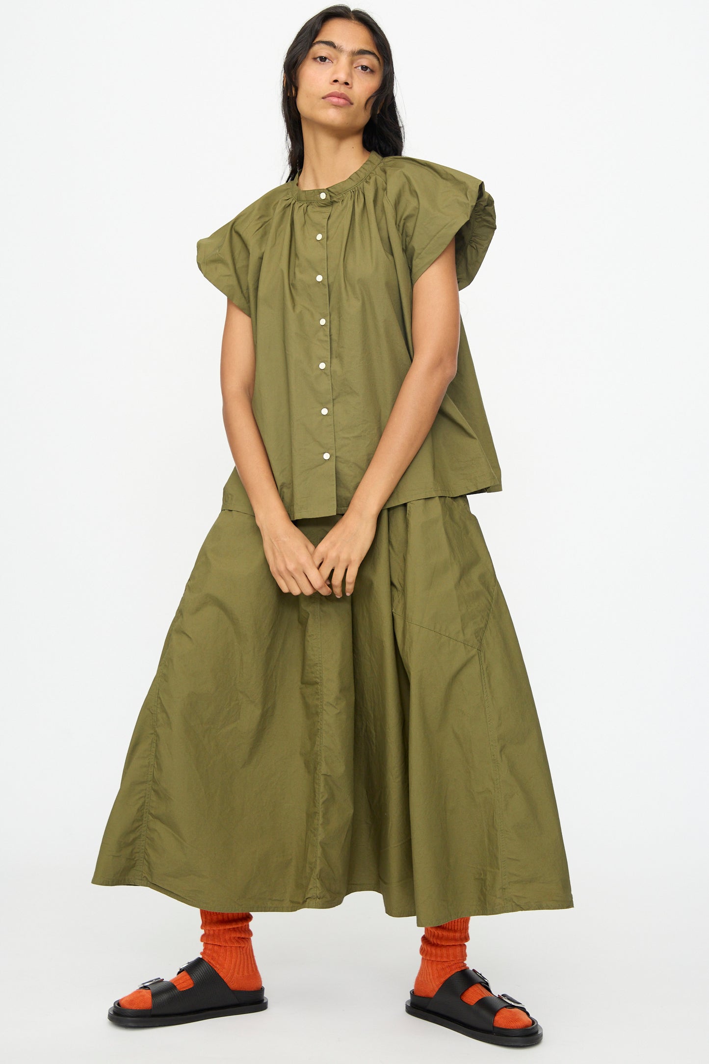 A woman stands against a plain background wearing an olive green short-sleeve button-up top, orange socks, black sandals, and the Ichi Antiquités Cotton Typewriter Skirt in Khaki.