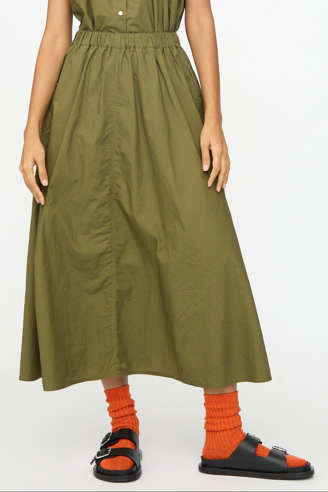 A person wears the Ichi Antiquités Cotton Typewriter Skirt in Khaki, paired with bright orange socks and black sandals, standing against a plain light background.