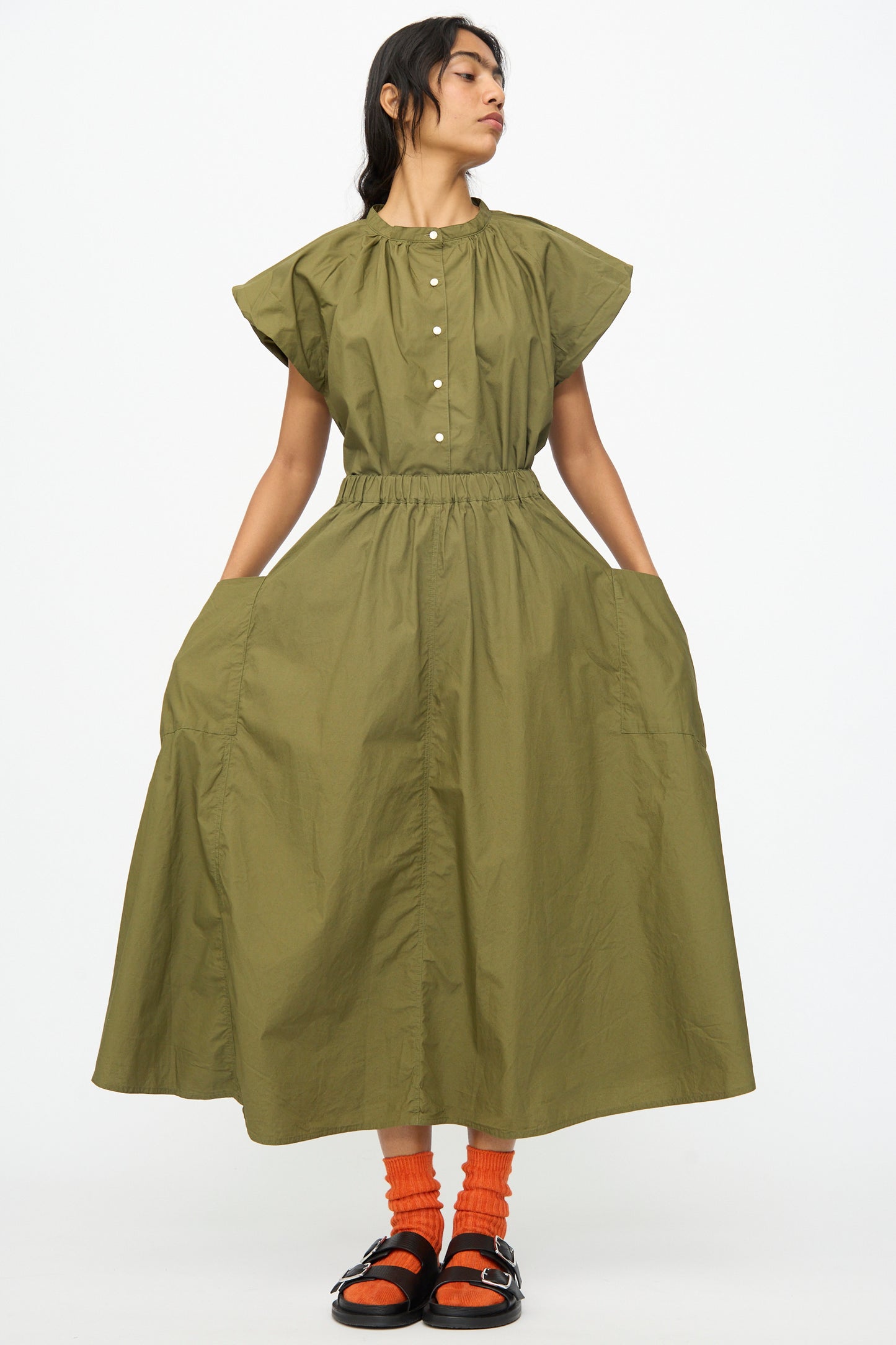 A woman stands against a plain white background wearing the Ichi Antiquités Cotton Typewriter Skirt in khaki, styled with orange socks and black sandals.