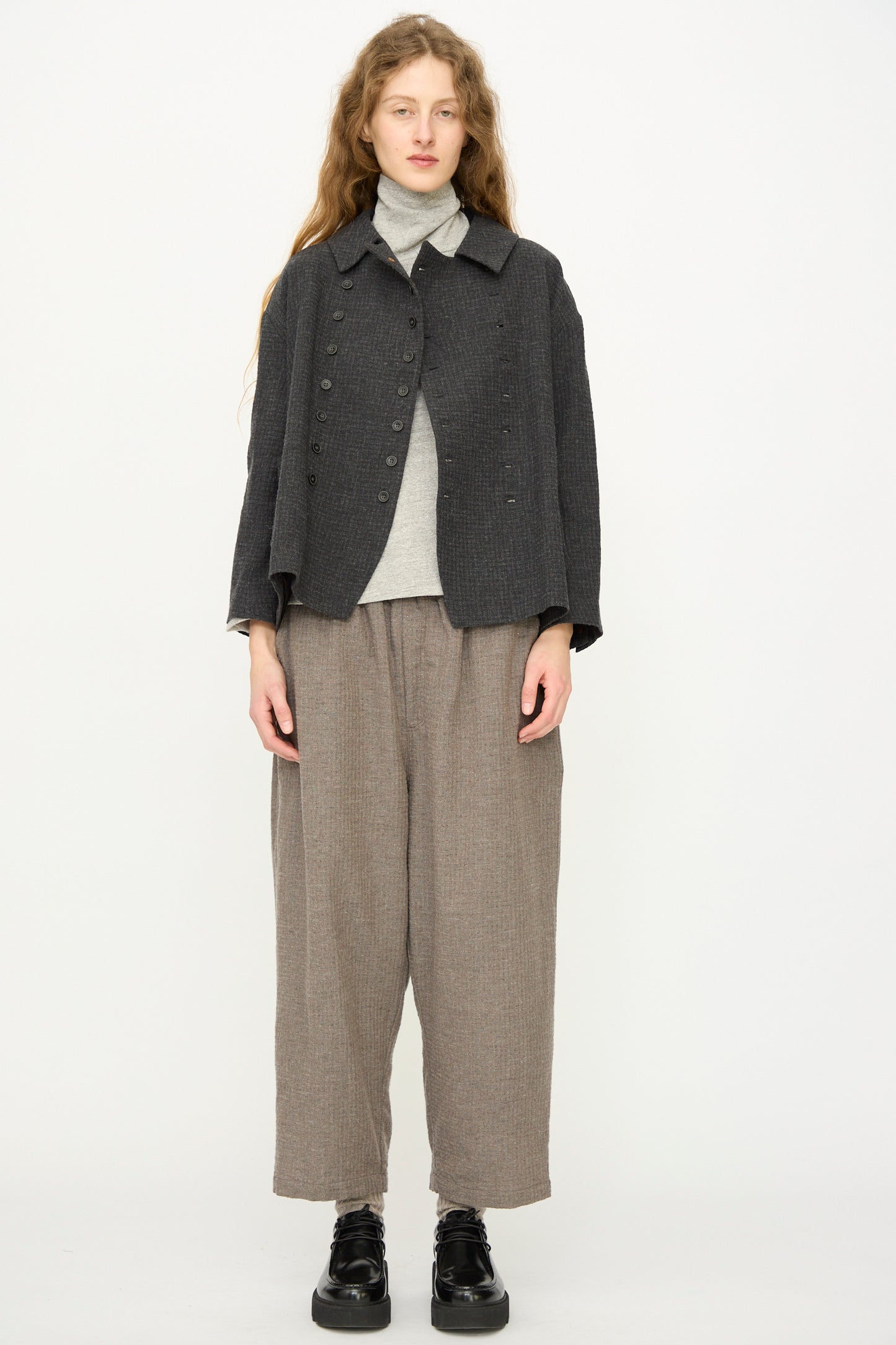 A woman with long hair wears the Ichi Antiquités Cotton Wool Sucker Jacket in Charcoal over a gray turtleneck and loose brown pants, standing against a plain white background.