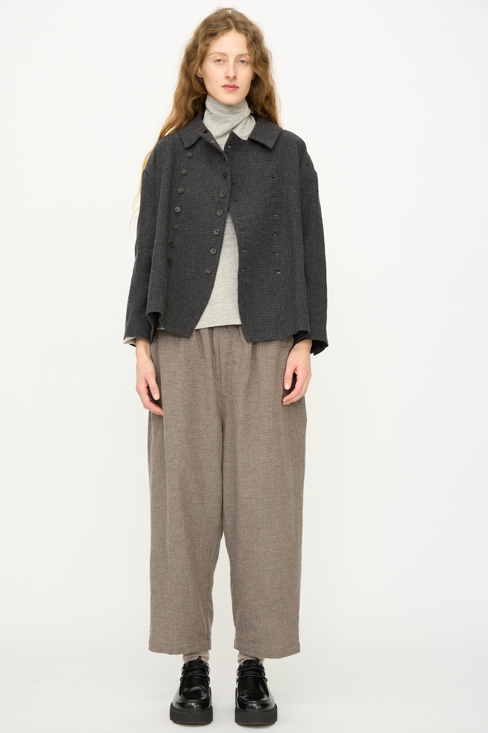 A woman with long hair wears the Ichi Antiquités Cotton Wool Sucker Jacket in Charcoal over a gray turtleneck and loose brown pants, standing against a plain white background.