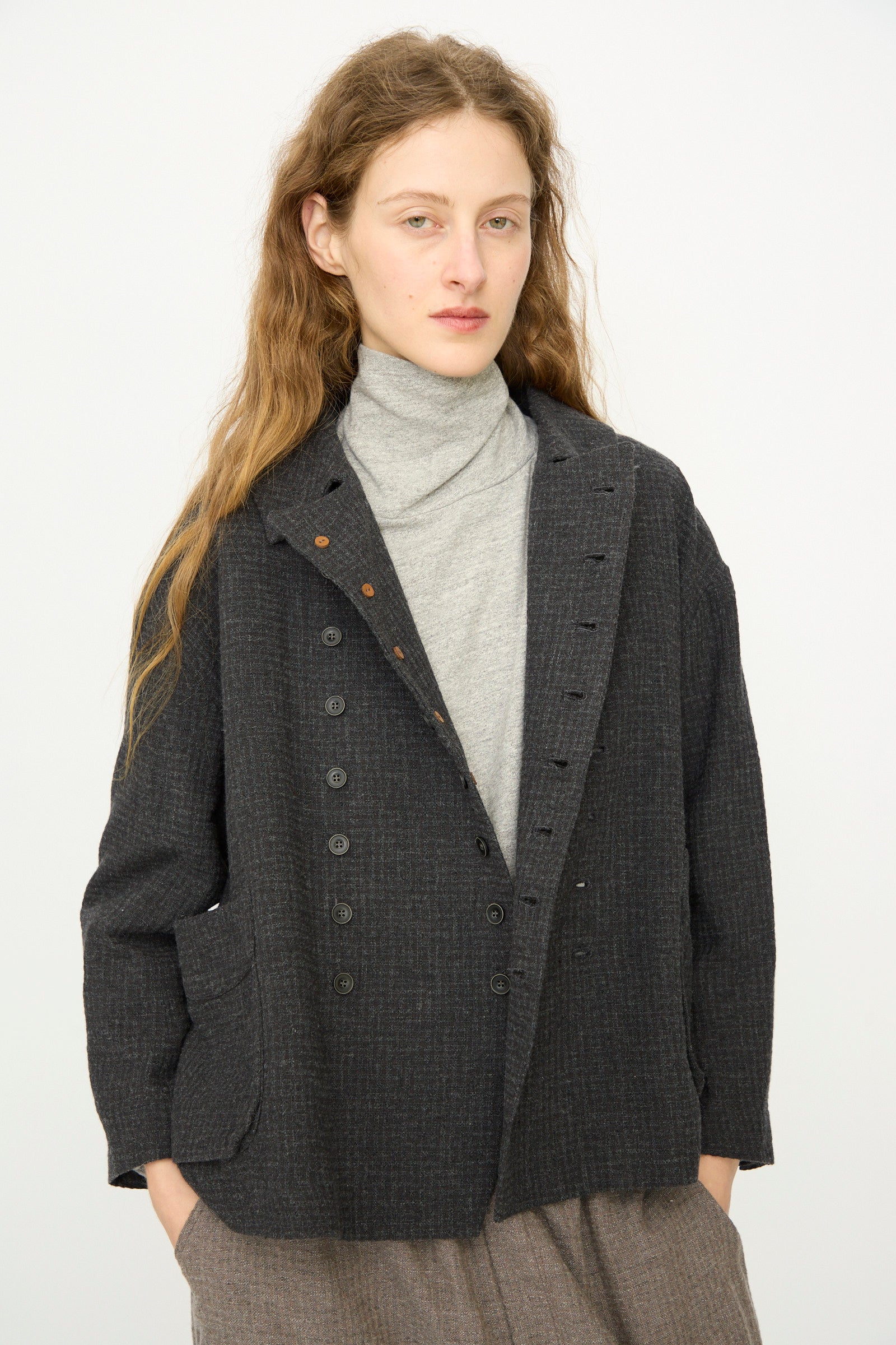 A person with long, wavy hair wears a gray turtleneck under the Ichi Antiquités Cotton Wool Sucker Jacket in Charcoal, styled with loose brown pants and standing against a plain light background.