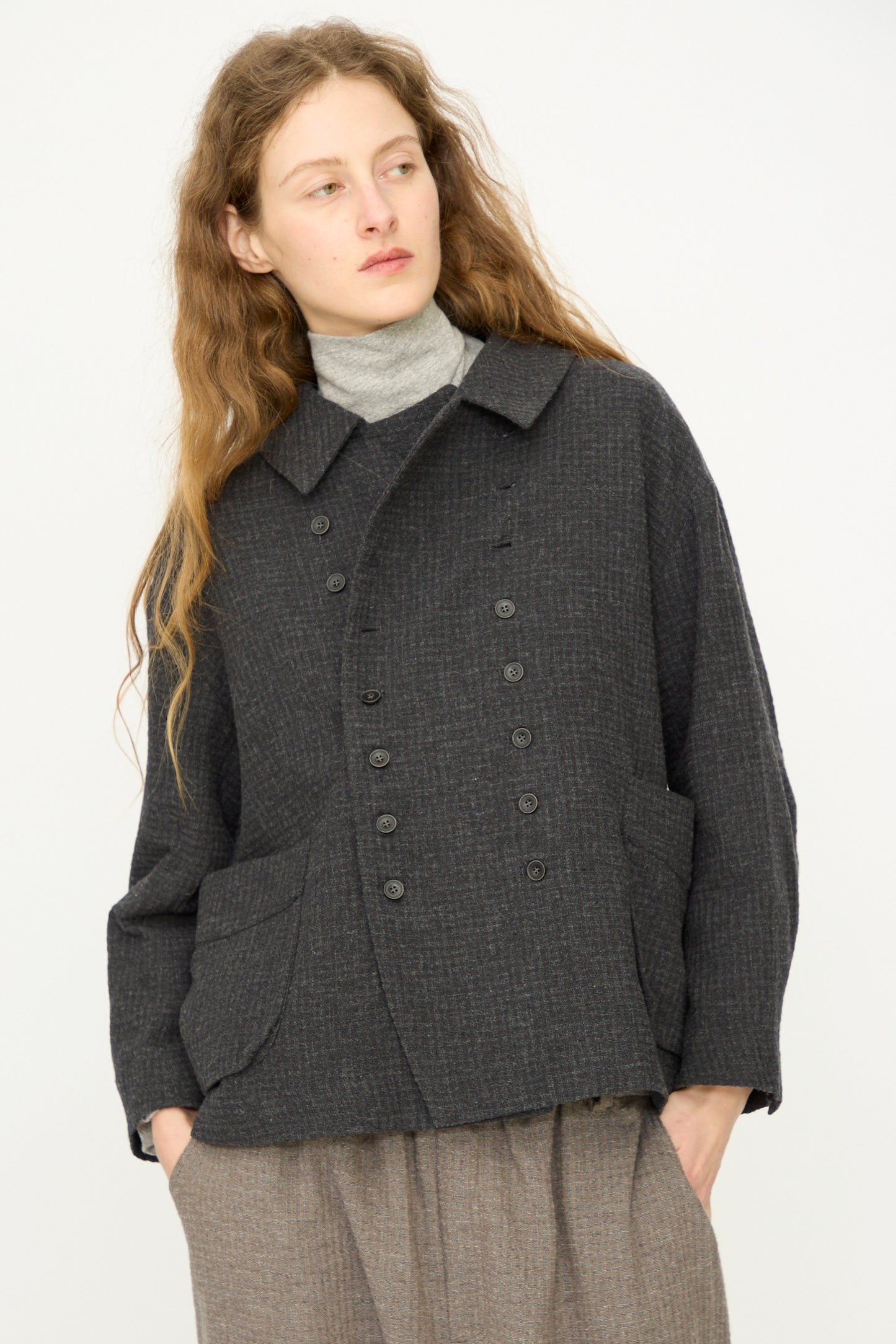 A person with long wavy hair wears the Ichi Antiquités Cotton Wool Sucker Jacket in Charcoal over a light turtleneck, hands in pockets, standing against a plain light background.