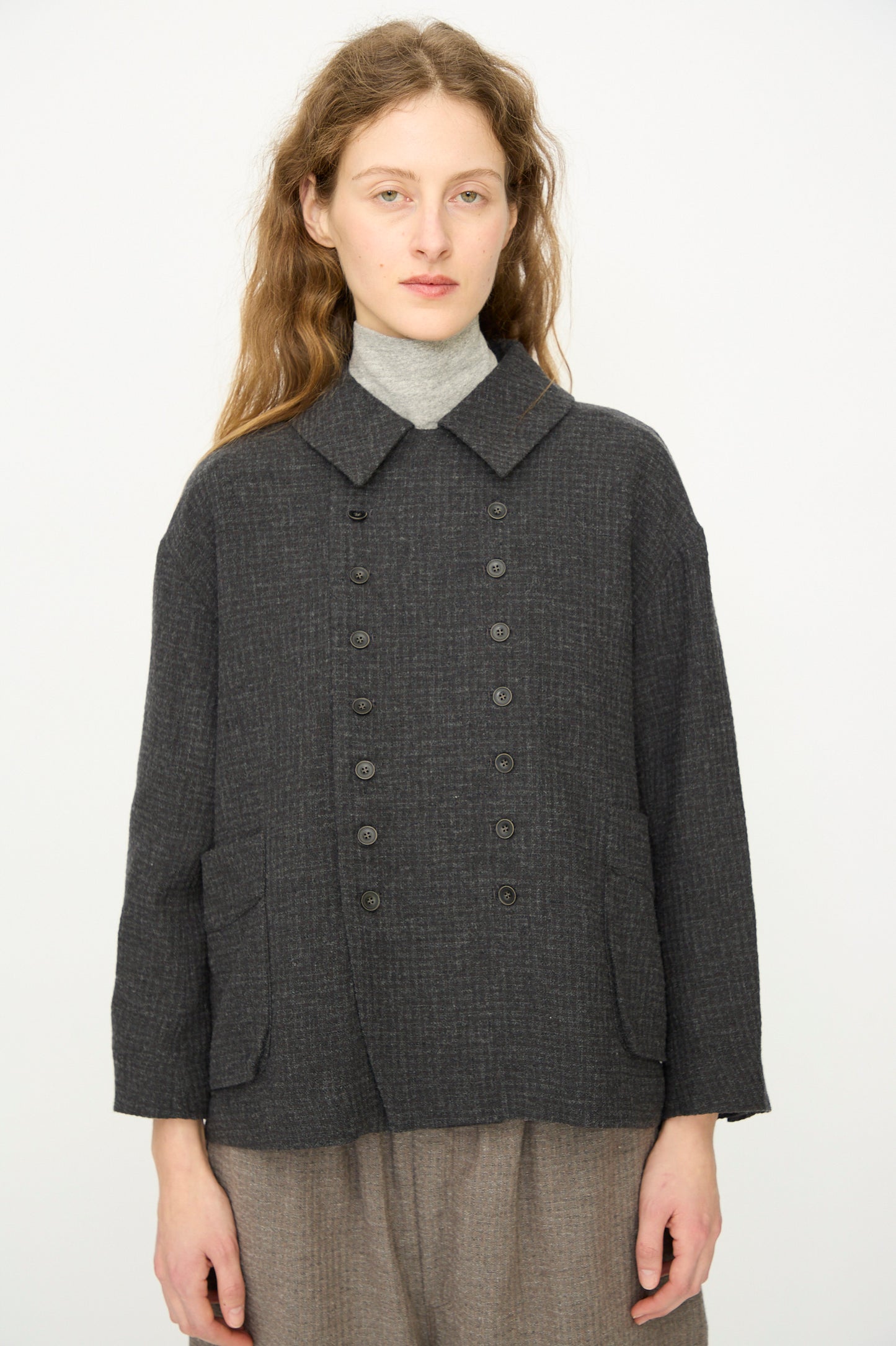 A person with long wavy hair wears a gray turtleneck, brown pants, and the Ichi Antiquités Cotton Wool Sucker Jacket in Charcoal, posing against a plain white background.