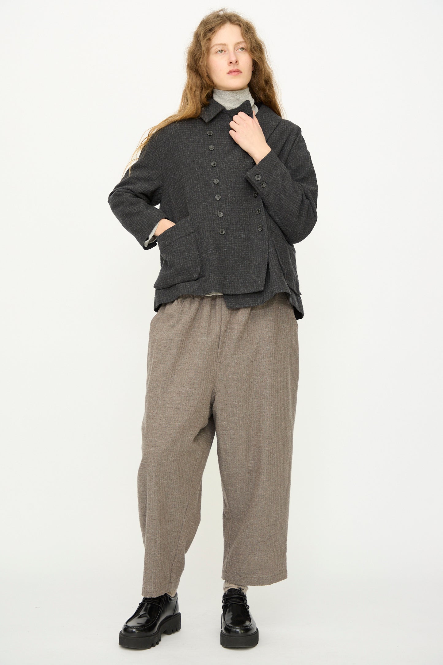 A person with long hair wears the Ichi Antiquités Cotton Wool Sucker Jacket in Charcoal over a light turtleneck and loose light brown pants, paired with black platform shoes, standing against a plain white background.