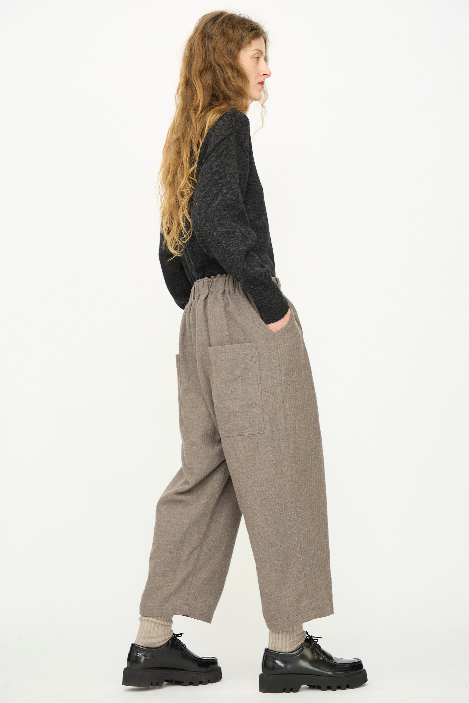 Person standing sideways against a plain background, wearing the Ichi Antiquités Cotton Wool Sucker Pant in Mocha with large pockets, a dark long-sleeve top, light socks, and black chunky shoes.