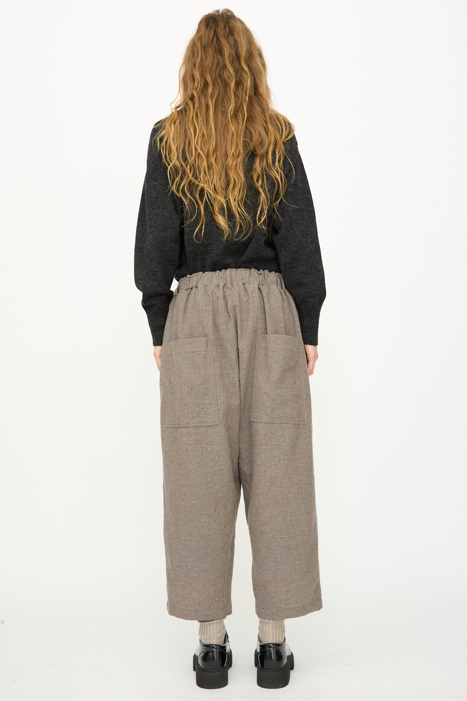 A person with long, wavy hair faces away, wearing a black sweater and Ichi Antiquités Cotton Wool Sucker Pant in Mocha, featuring a relaxed wide-leg fit and large back pockets, plus gray socks and black platform shoes against a white background.