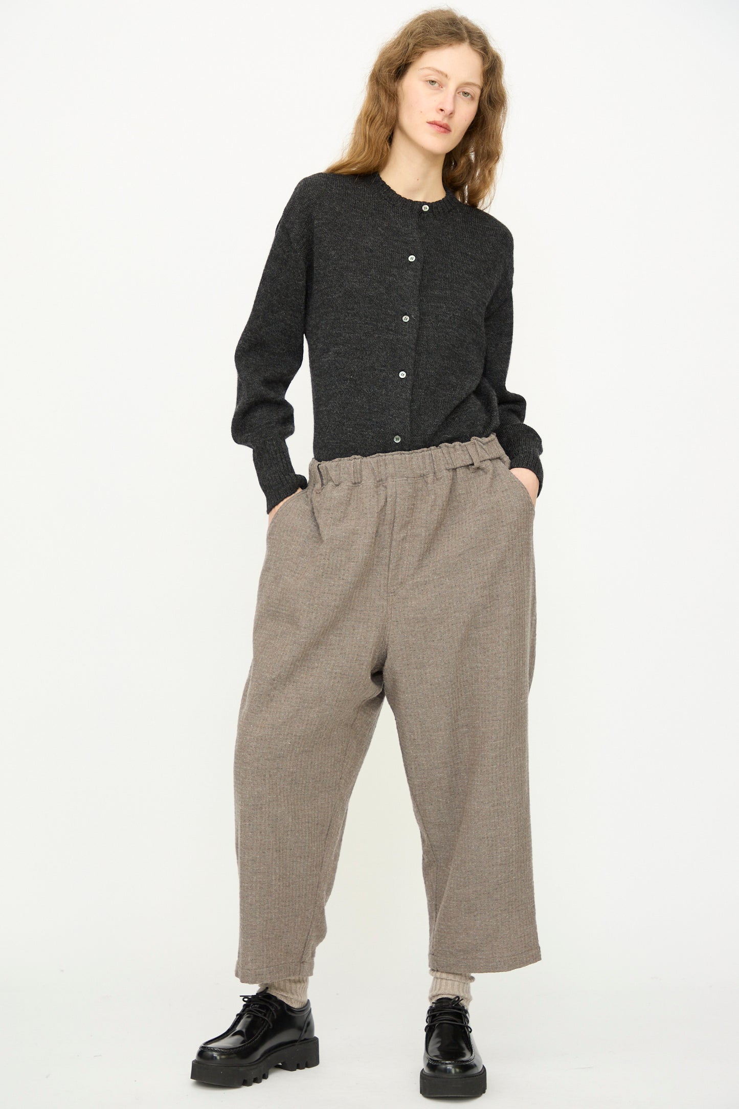 Person stands against a plain background, wearing a dark buttoned sweater, Ichi Antiquités Cotton Wool Sucker Pant in Mocha with relaxed wide-leg fit, and black shoes, hands in pockets.
