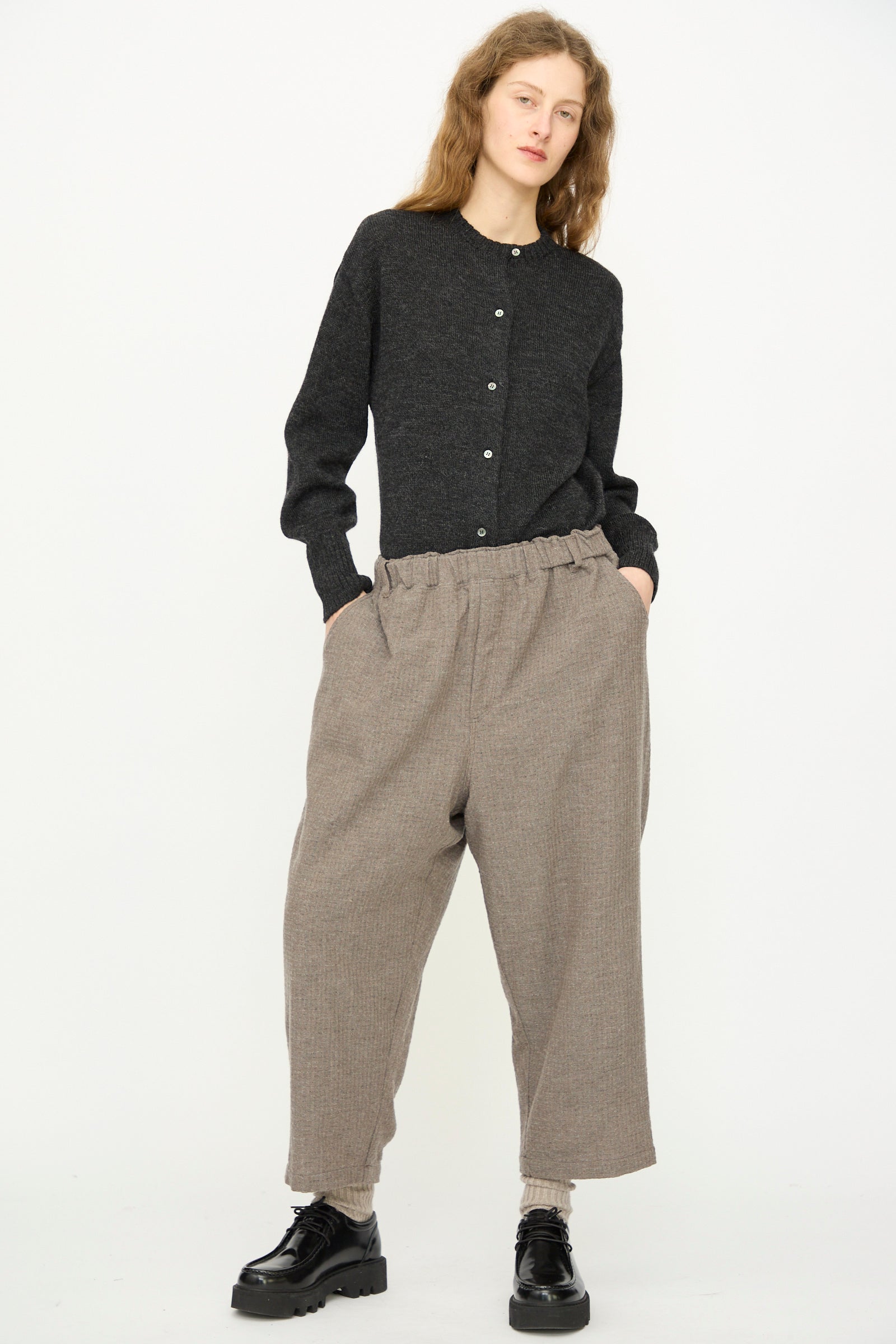 Person stands against a plain background, wearing a dark buttoned sweater, Ichi Antiquités Cotton Wool Sucker Pant in Mocha with relaxed wide-leg fit, and black shoes, hands in pockets.