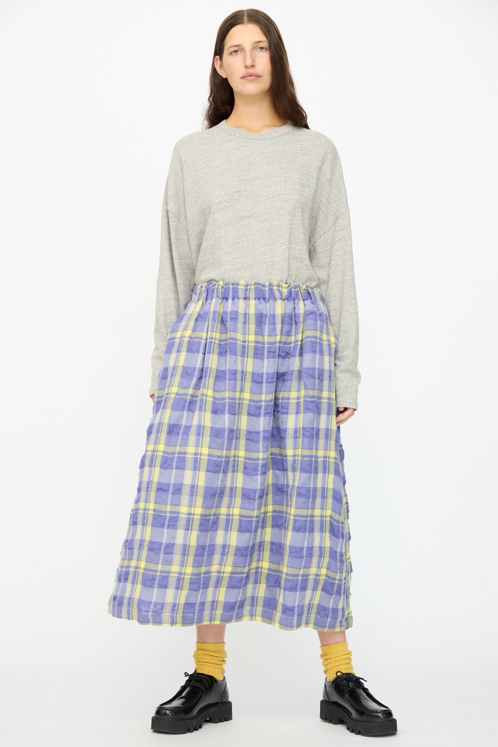 A person stands facing forward against a white background, wearing a gray long-sleeve top, Ichi Antiquités’ Cotton Wool Tartan Check Skirt in Blue with an elastic waist, yellow socks, and black platform shoes.