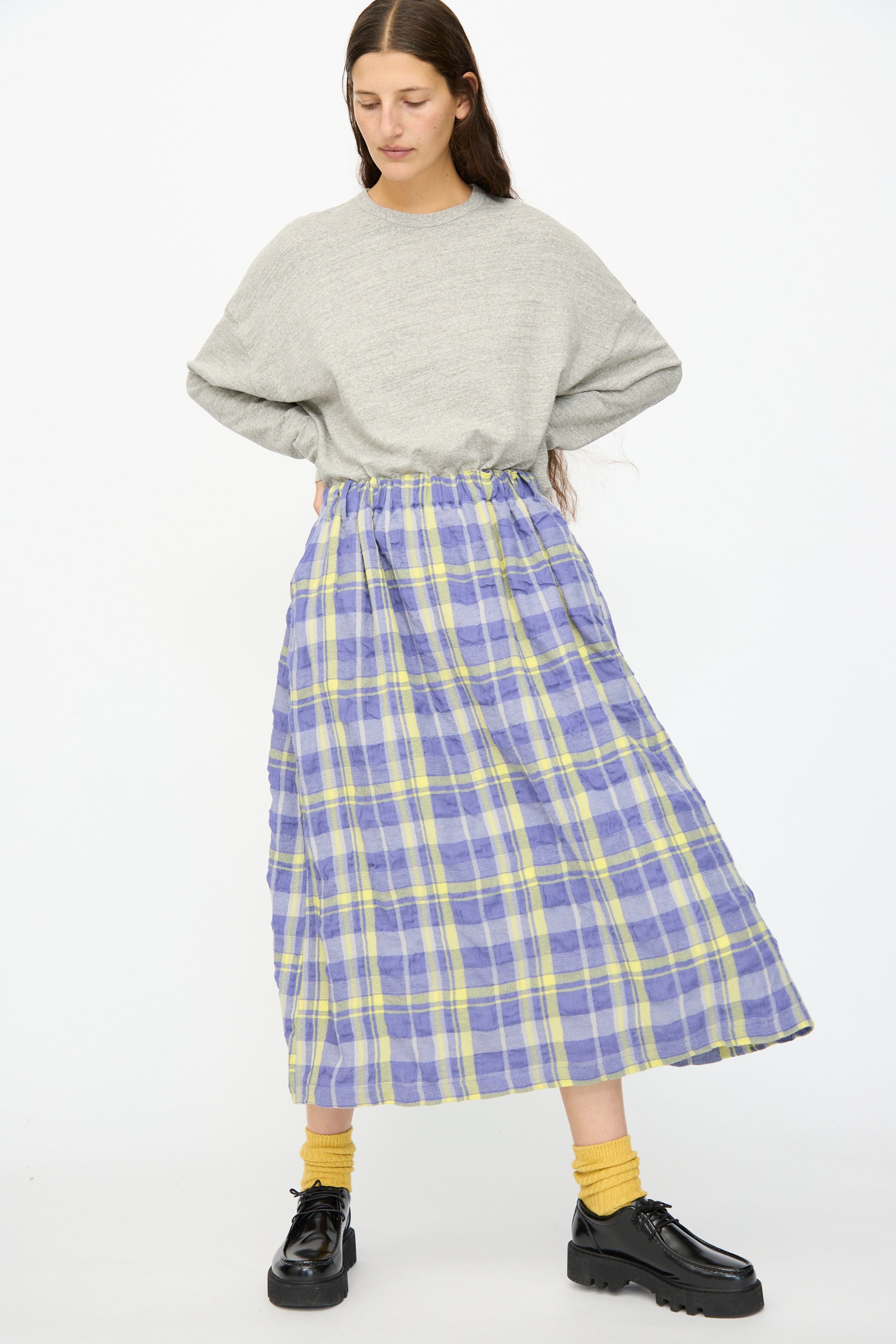 A person stands with hands on hips, wearing a loose grey sweater, yellow socks, black shoes, and the Ichi Antiquités Cotton Wool Tartan Check Skirt in Blue against a plain white background.