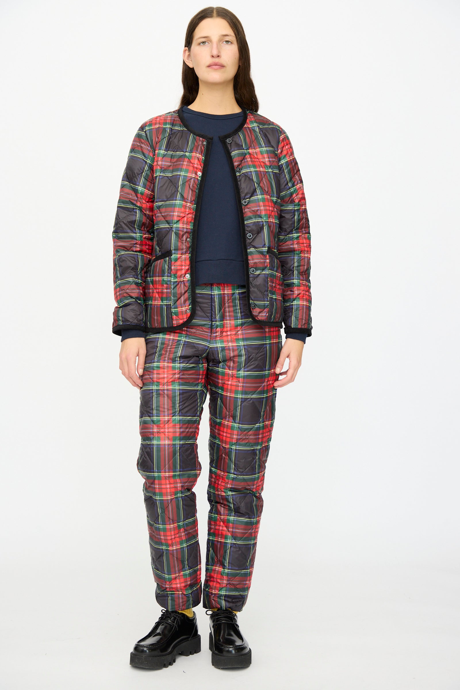 A person faces forward against a plain white background, wearing the Ichi Antiquités Down Jacket in Black Tartan over a navy top, paired with matching pants featuring black corduroy trim and black shoes.
