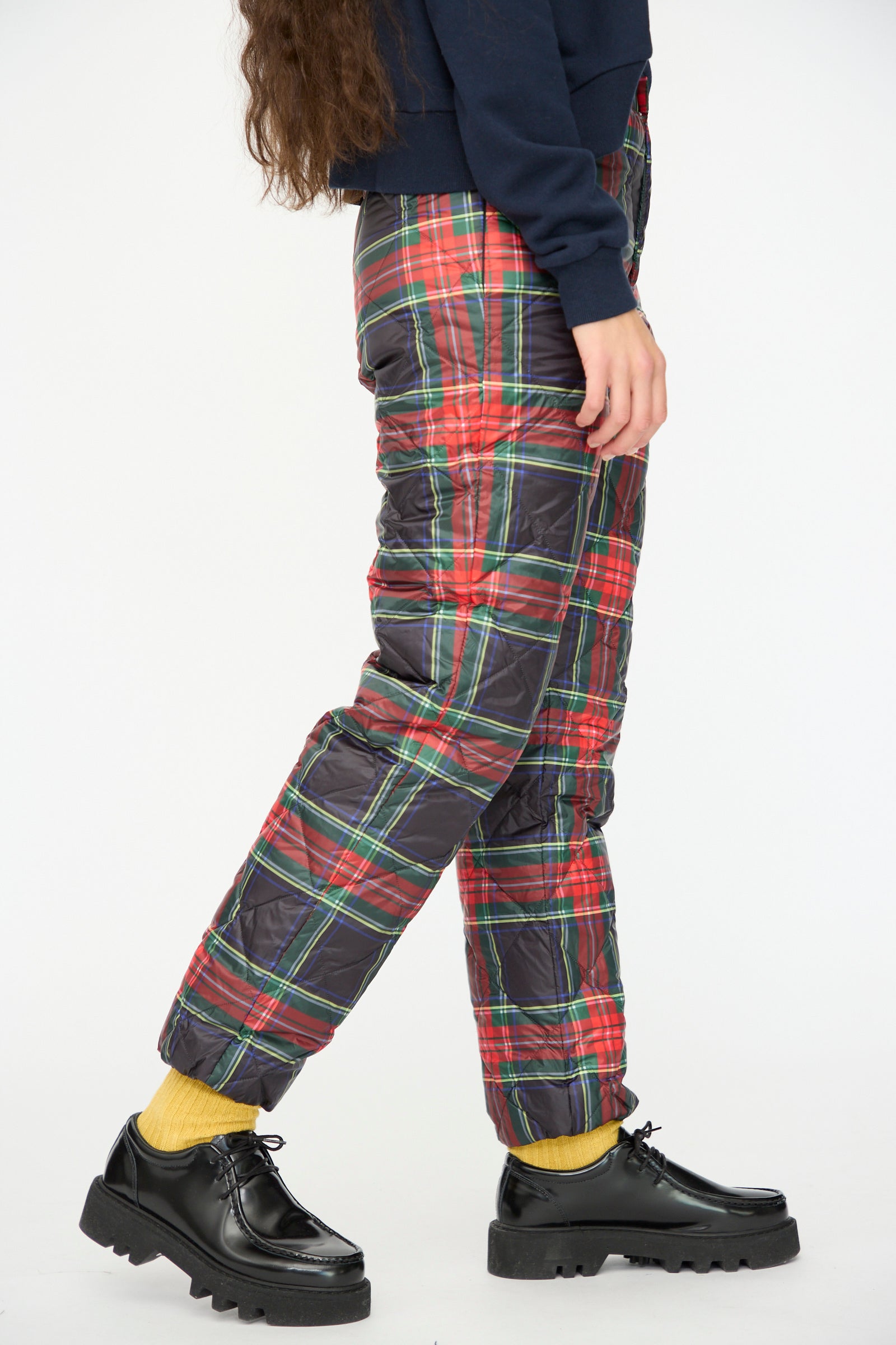 A person wearing Ichi Antiquités Down Pants in Black Tartan with an elastic waist, yellow socks, black platform shoes, and a dark top stands against a plain white background.