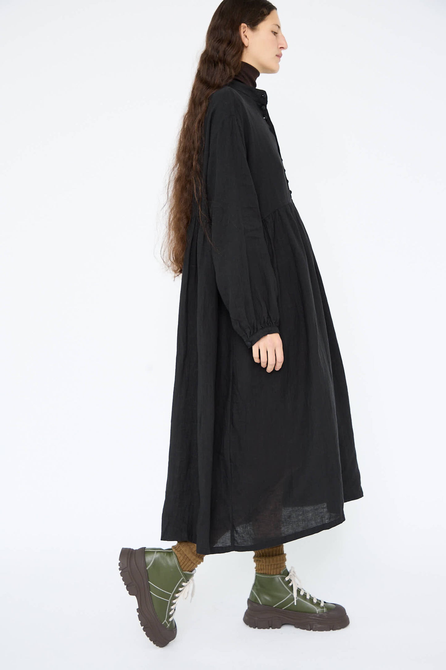 A person in a relaxed-fit, long French Linen Dress in black from Ichi Antiquités and green boots walks sideways against a white background.