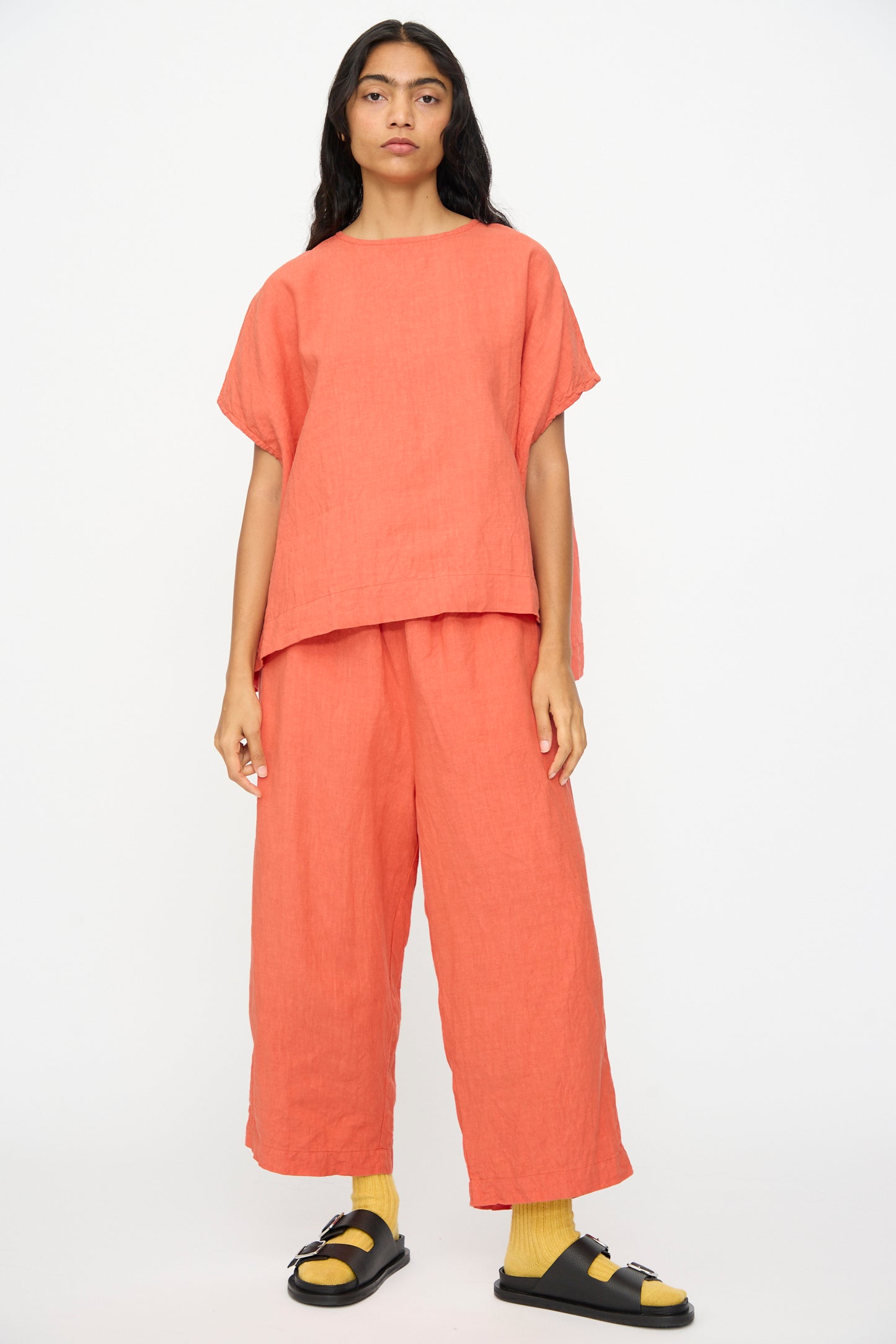 A person wears a loose coral top and Ichi Antiquités French Linen Pants in Orange—elastic waist, wide cropped legs—paired with yellow socks and black sandals, standing against a plain white background.