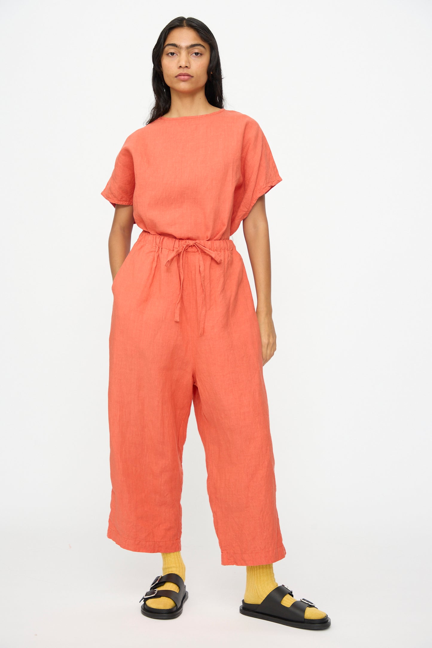 A person models Ichi Antiquités French Linen Pants in orange, featuring an elastic waistband, paired with yellow socks and black sandals, against a plain white background.