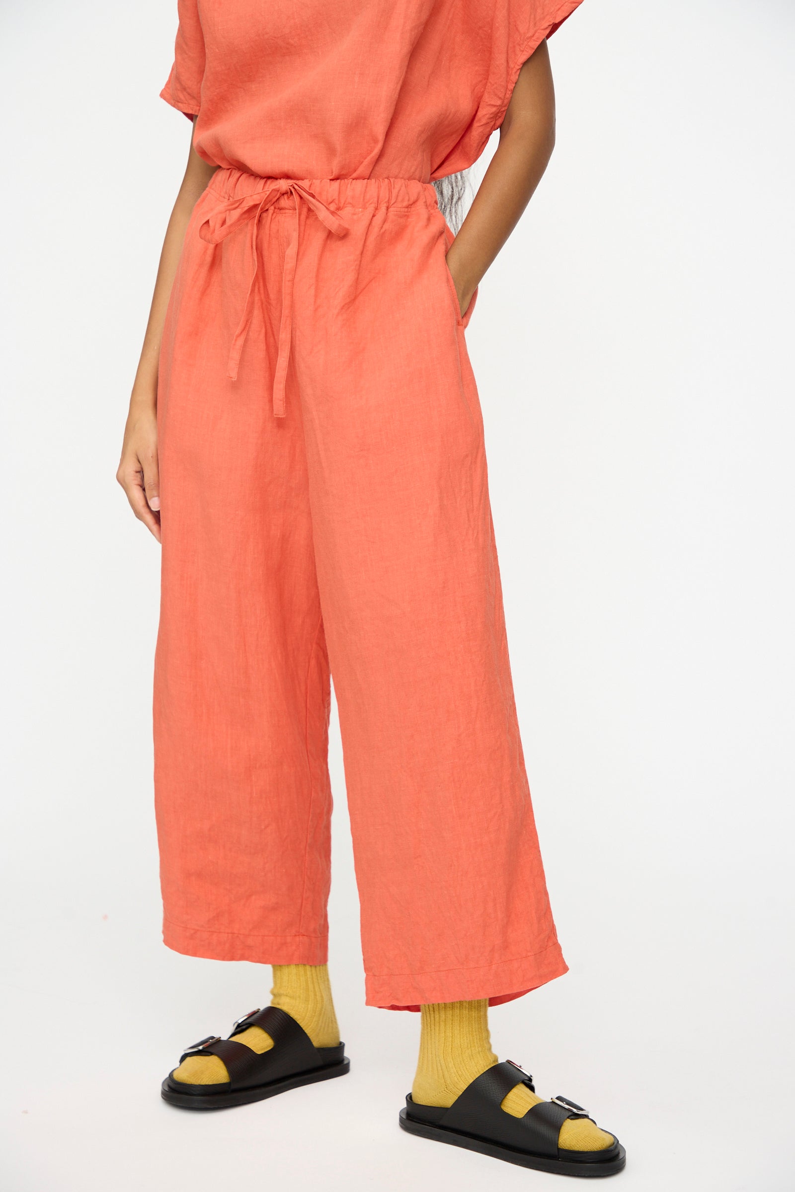 A person wears Ichi Antiquités French Linen Pants in Orange, featuring an elastic waistband and wide cropped leg, styled with yellow socks and black sandals, standing against a plain white background.