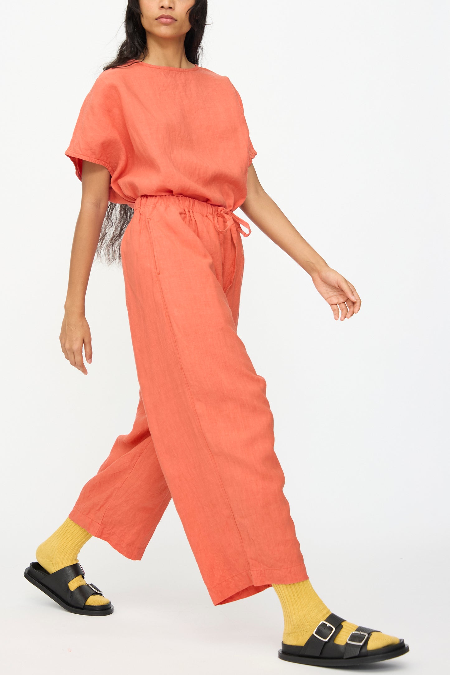 Someone wearing Ichi Antiquités French Linen Pants in Orange, featuring a wide cropped leg and elastic waistband, pairs them with yellow socks and black sandals while walking against a plain white background.