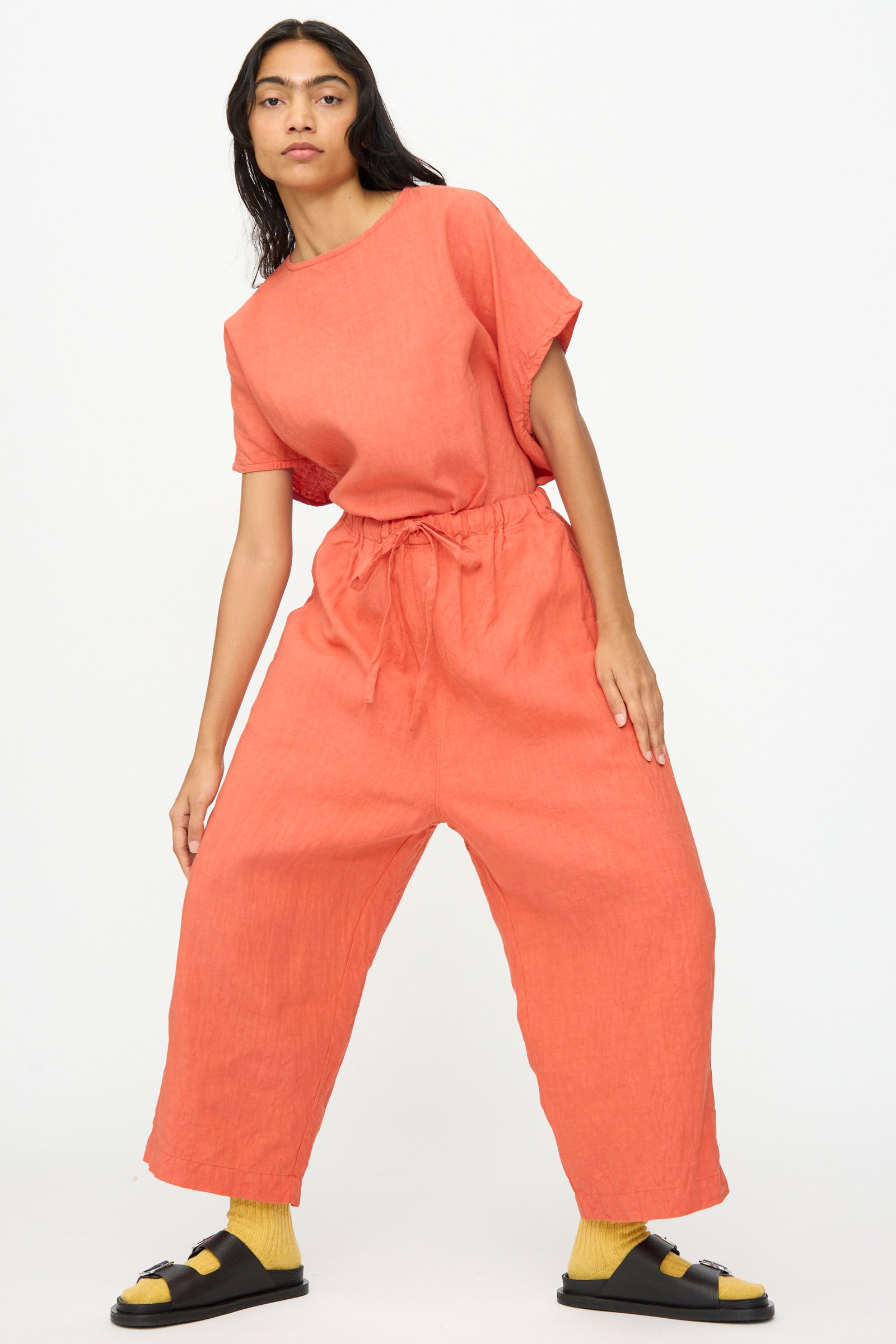 A woman stands in a relaxed pose wearing Ichi Antiquités French Linen Pants in orange, paired with yellow socks and black sandals, against a plain white background.