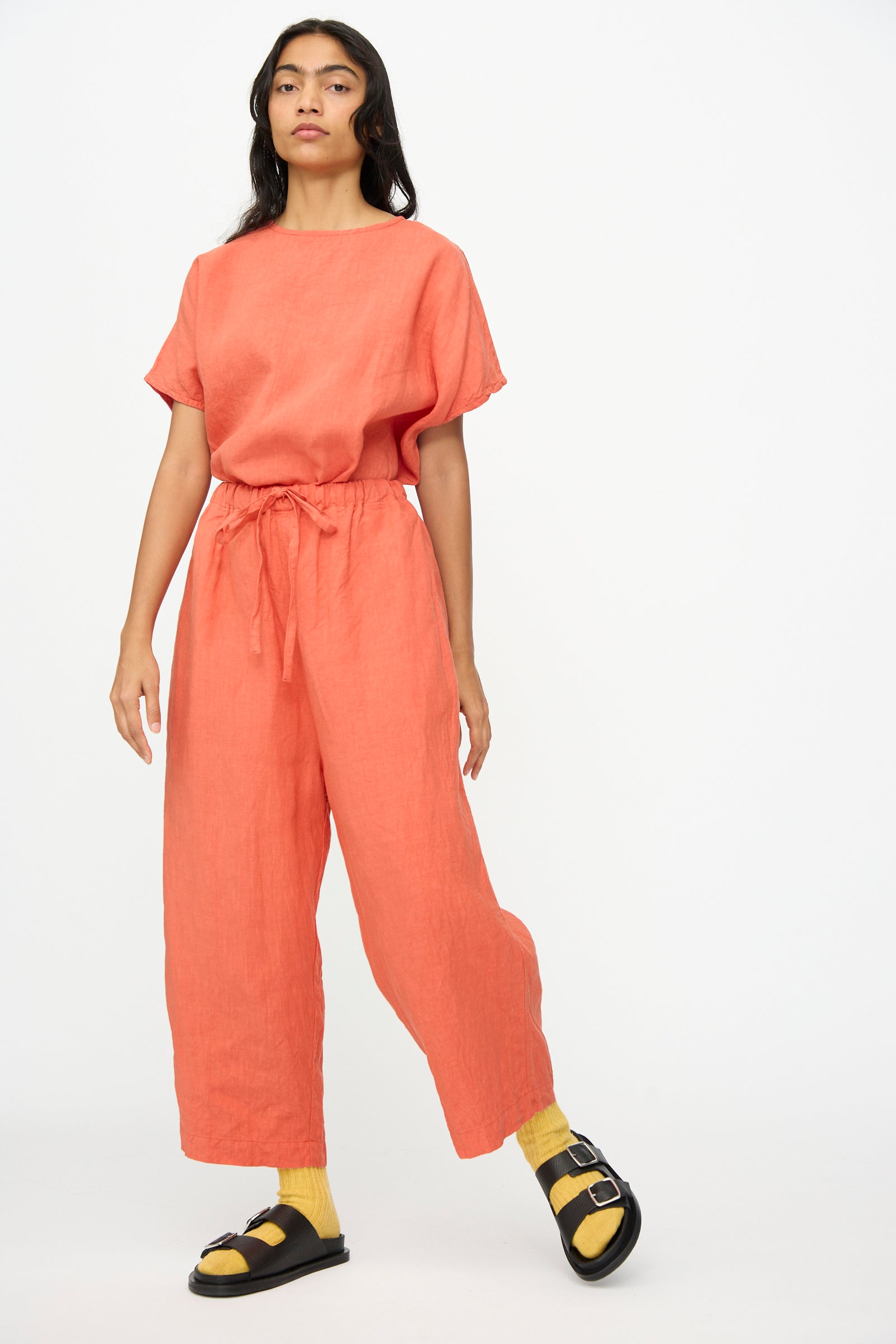 Wearing Ichi Antiquités' French Linen Pants in Orange—wide, cropped, with an elastic waist—paired with a loose coral short-sleeve top, yellow socks, and black sandals, this person stands against a plain white background.
